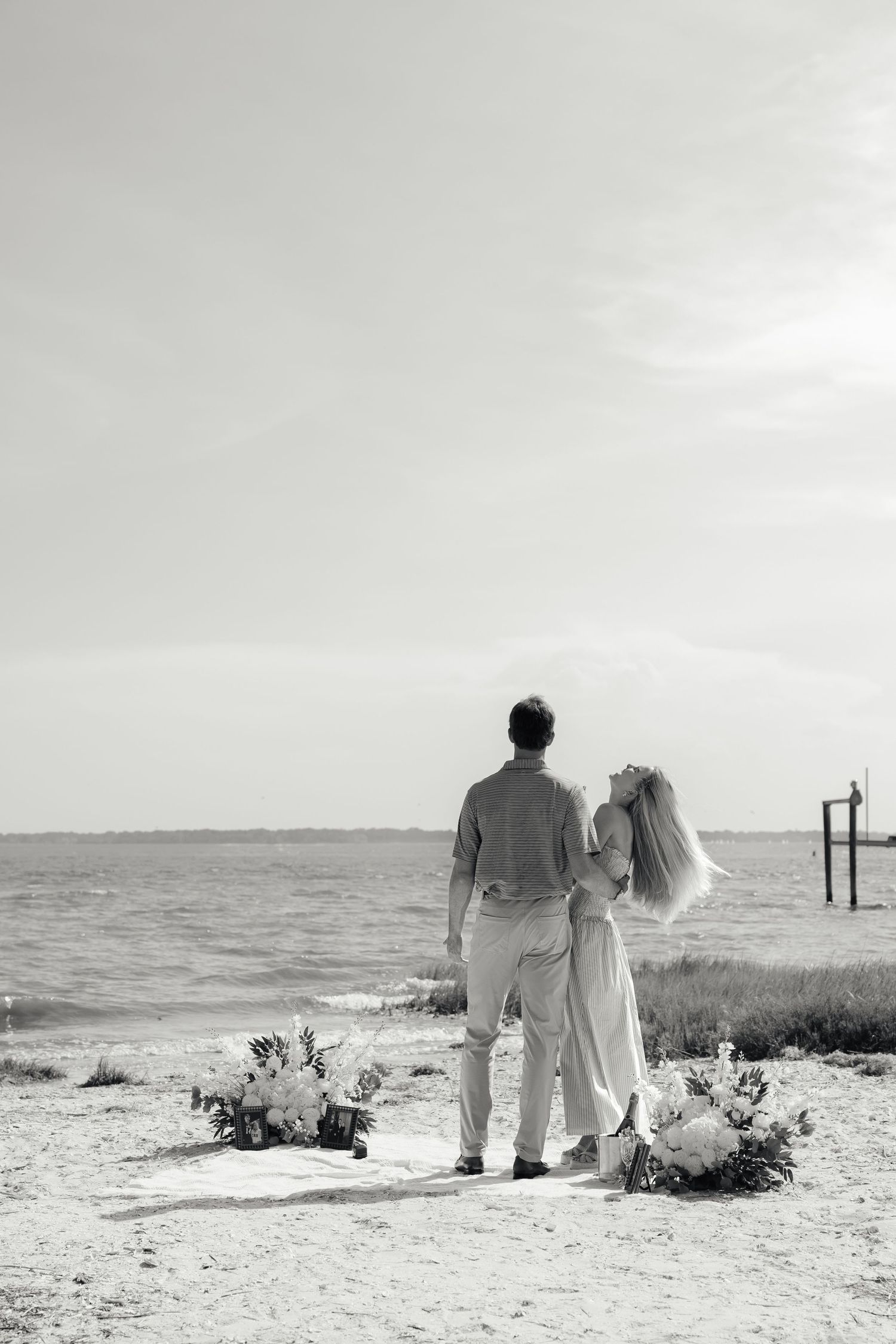 Black and white photo of couple standing together gazing at ocean from sandy shore.