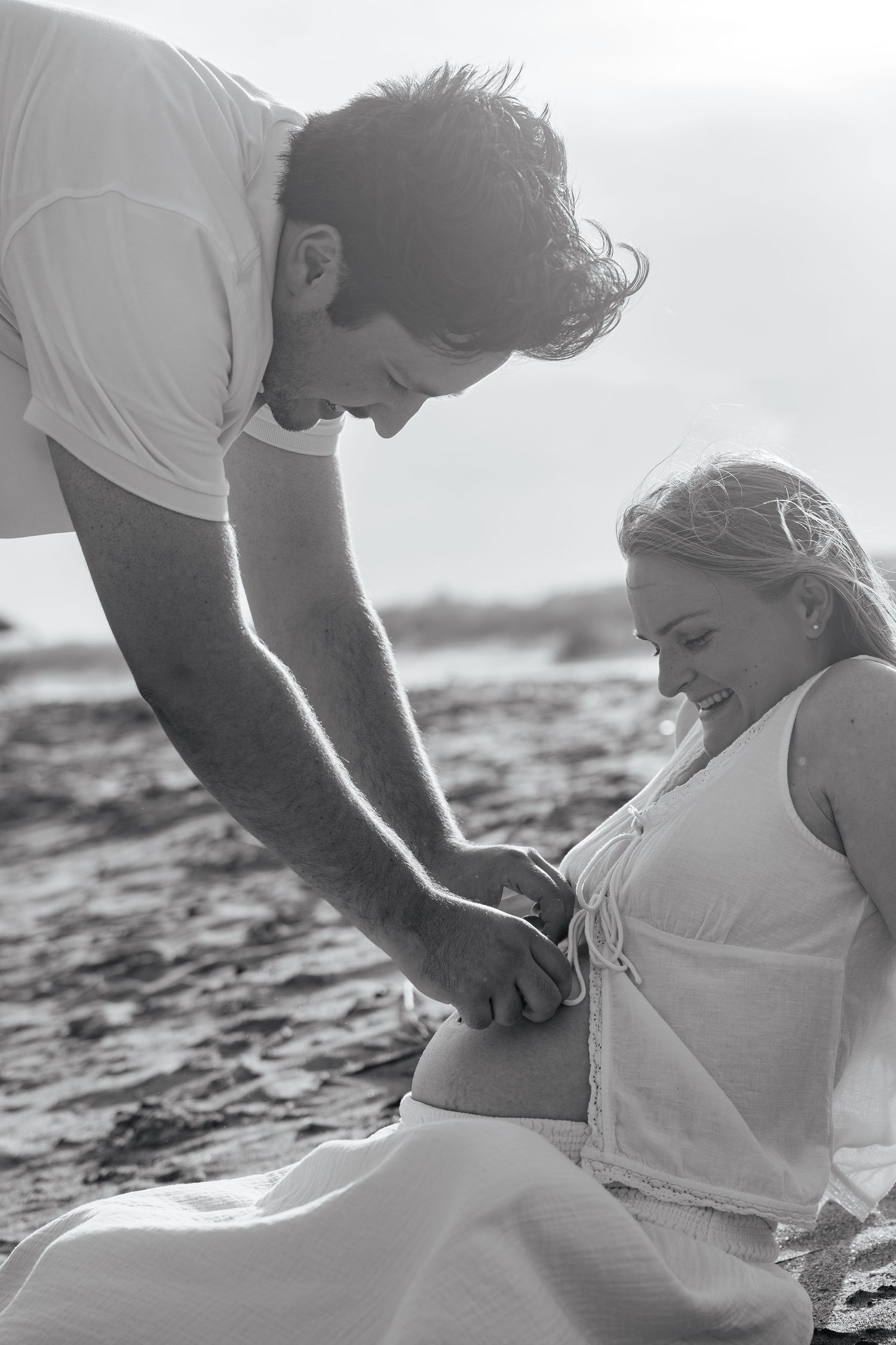 Black and white image of a man touching a pregnant woman's baby bump on the beach.