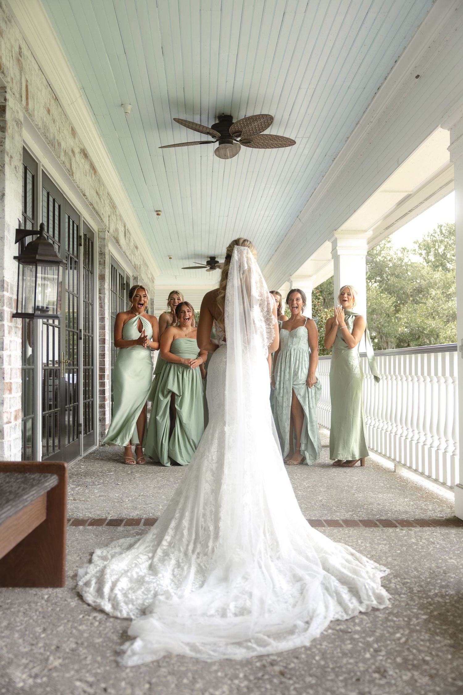 Bride in white lace gown and veil faces bridesmaids in sage green dresses on a porch during first look.