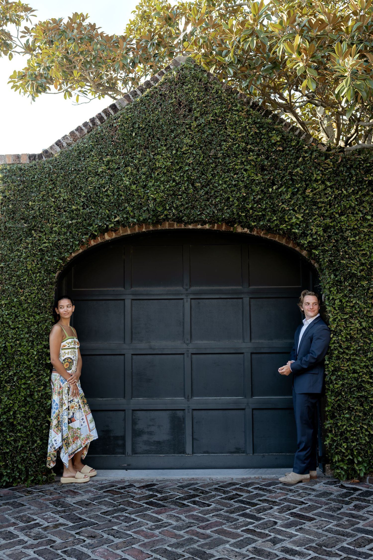 Engaged couple standing apart by an ivy-covered arched garage door on a brick driveway.