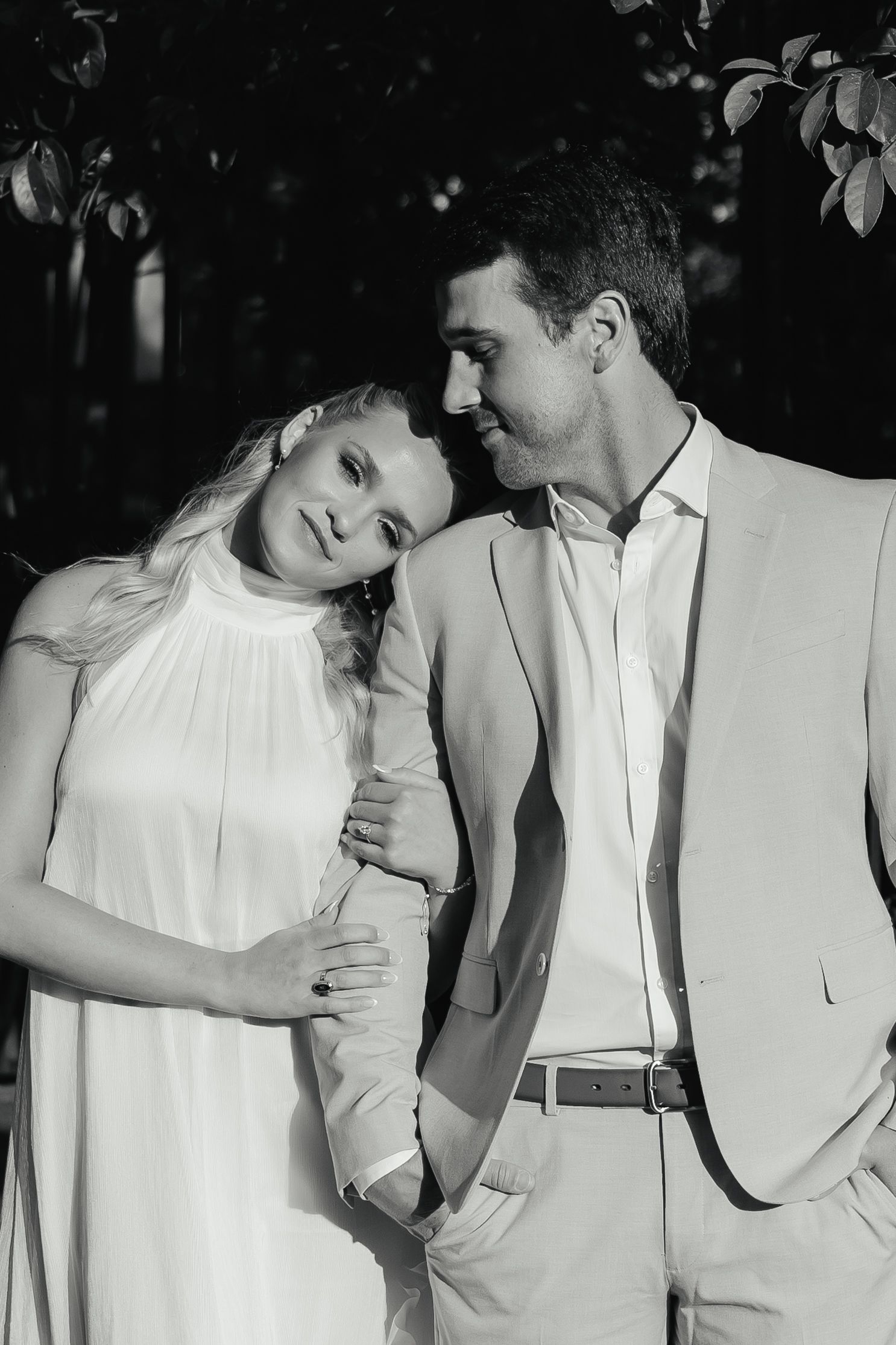 Black and white romantic portrait of couple standing close together under leafy trees outdoors.