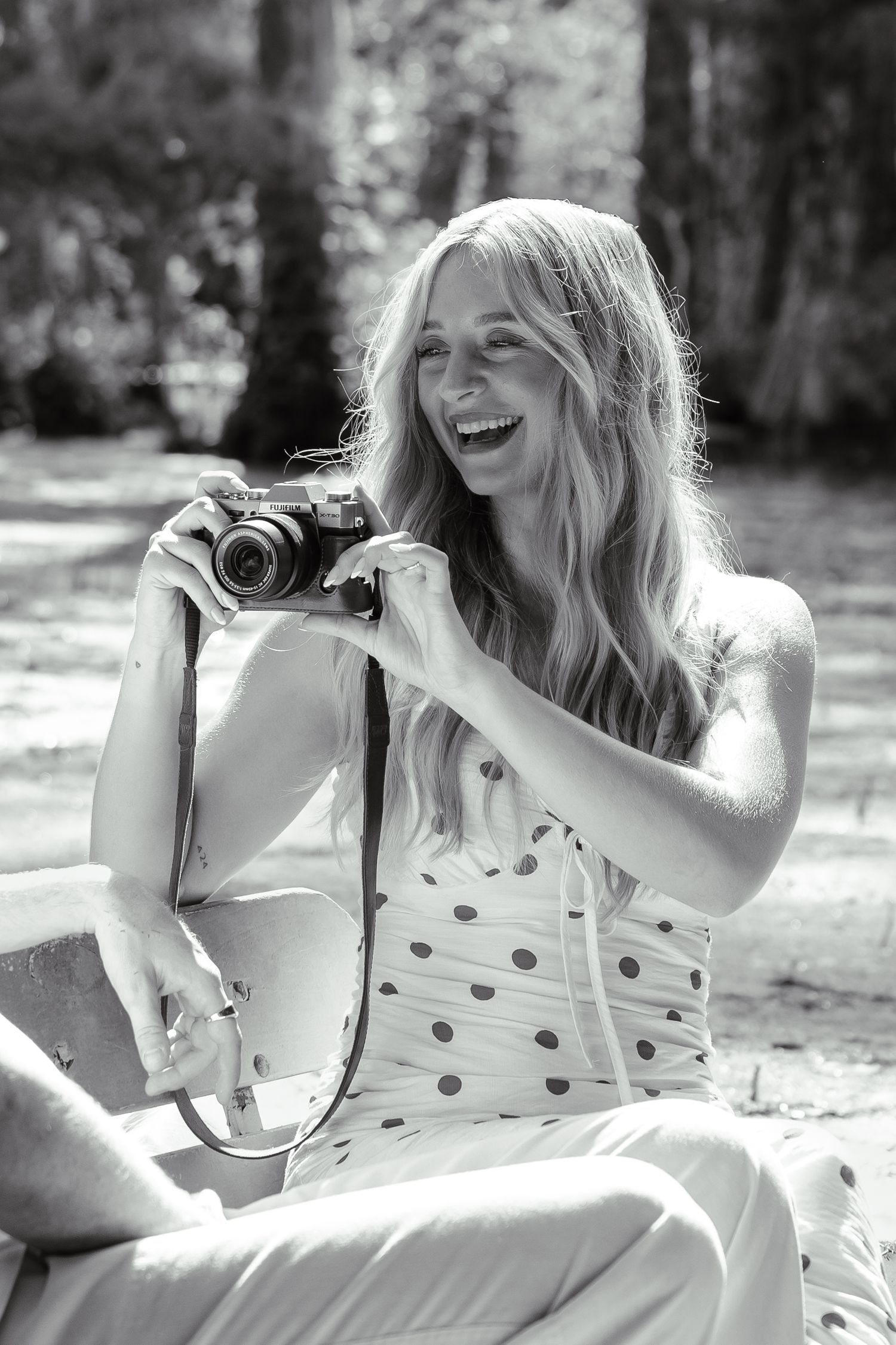 Black and white portrait of a laughing blonde woman holding a vintage camera outdoors.