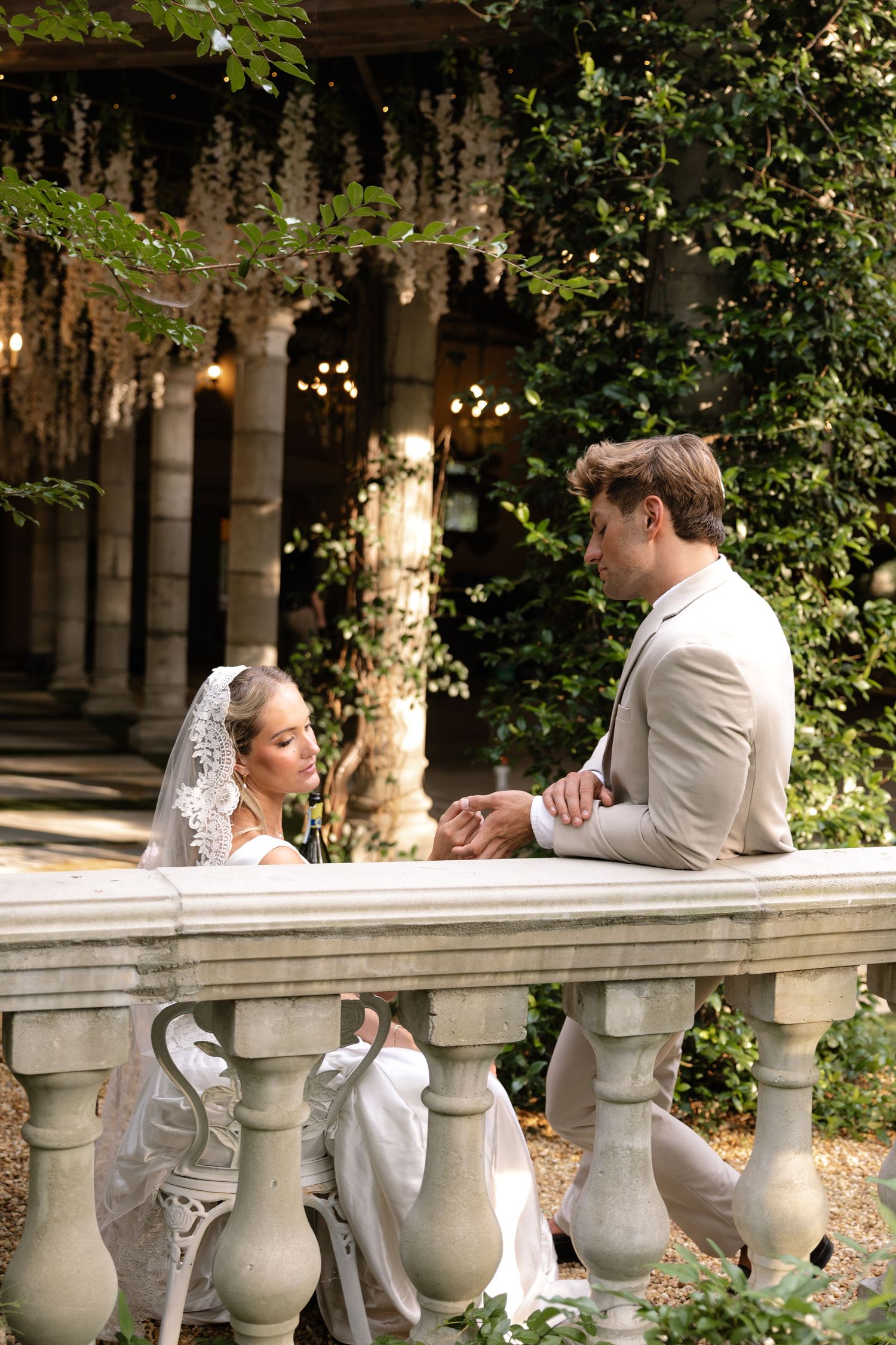 Bride and groom holding hands at a stone balustrade in a lush garden wedding venue with hanging floral decor.