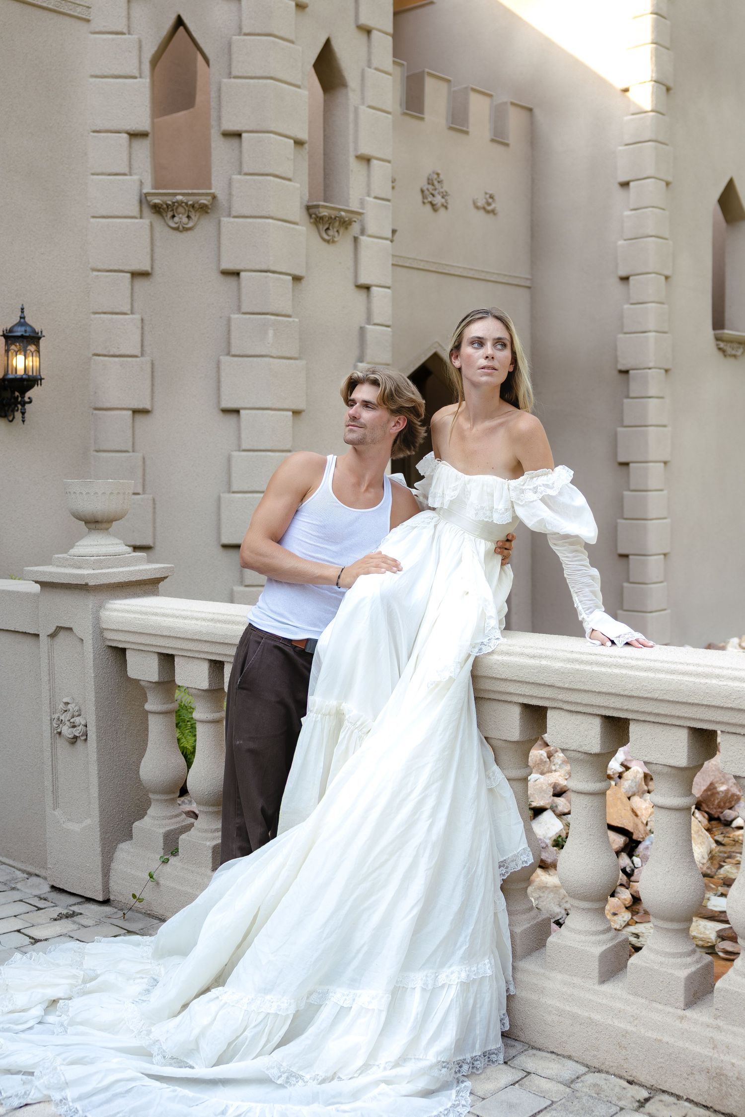 Couple posing on castle balcony, woman in elegant white off-shoulder wedding gown with long train.