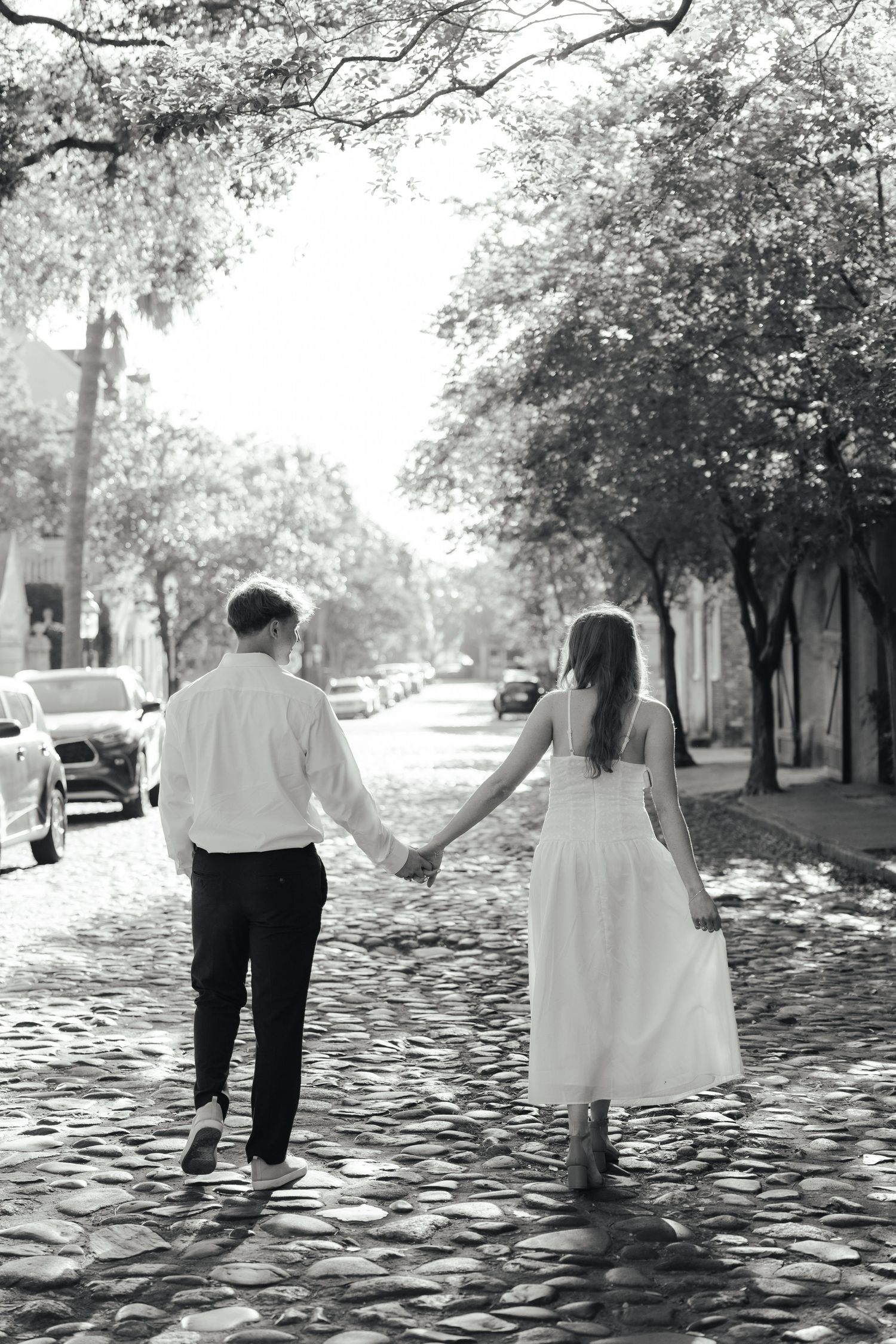 Black and white photo of a couple holding hands walking on a cobblestone street lined with trees and parked cars.