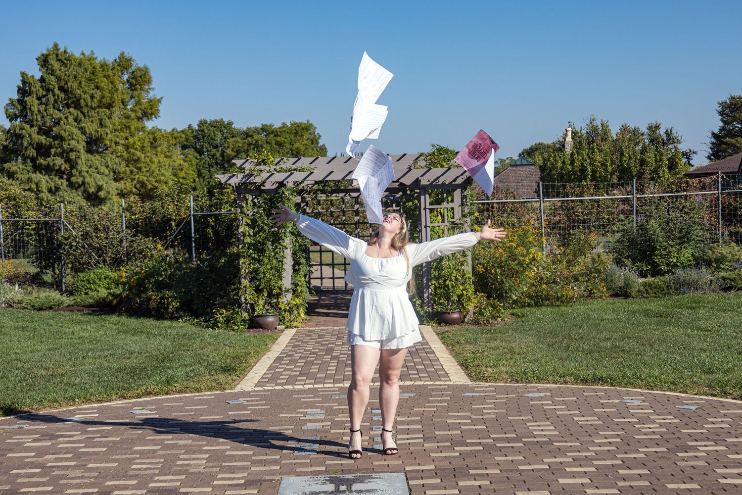 Person in white clothing joyfully throws papers in the air while standing on a brick patio.