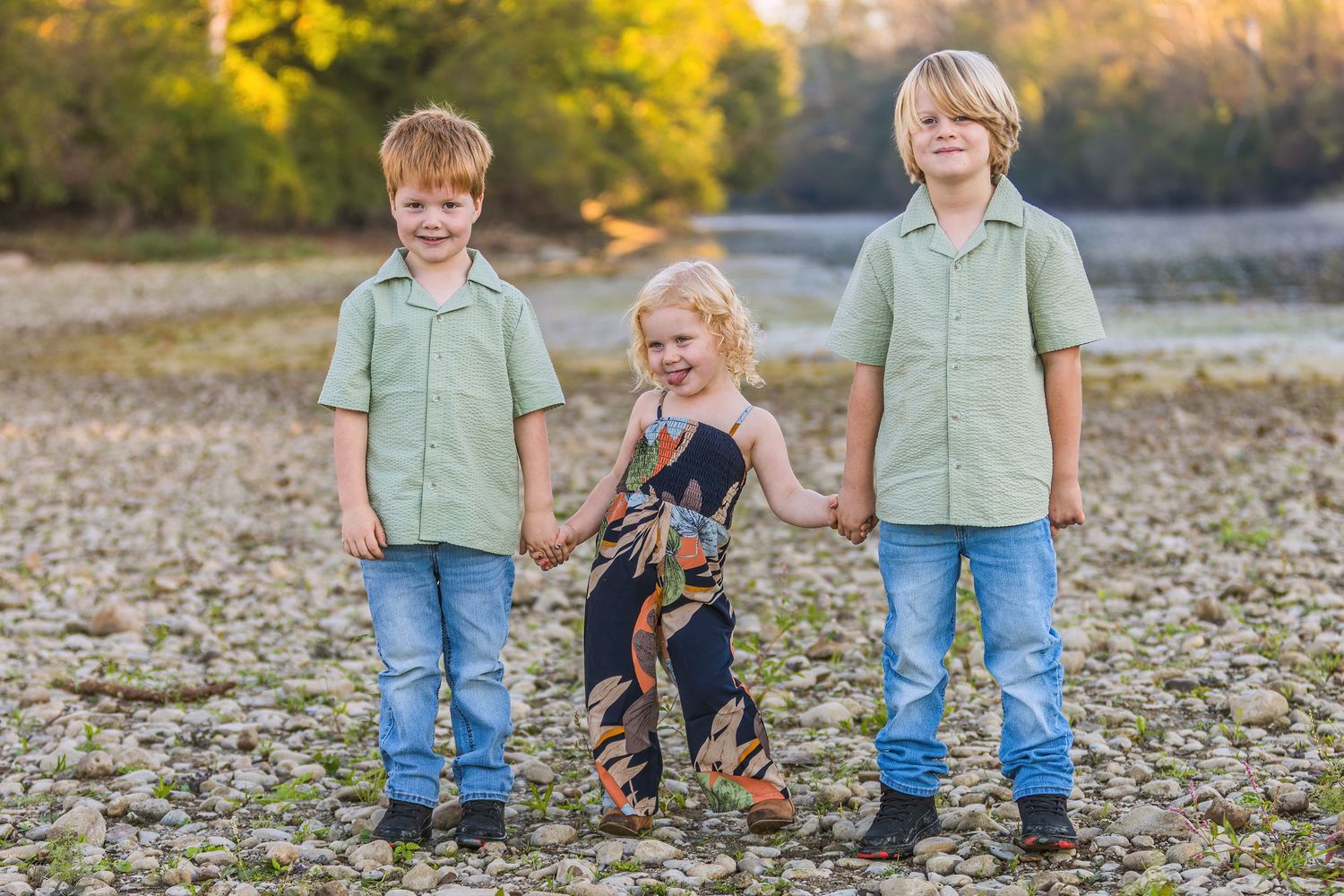 Three children in matching green shirts stand together on rocky ground with autumn foliage in the background.