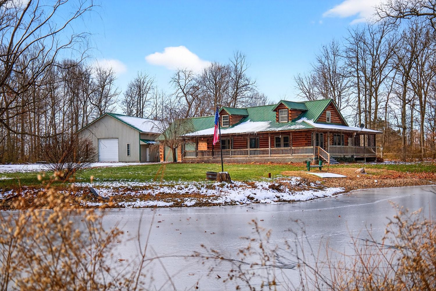 Rustic log cabin home with wrap-around porch sits beside a frozen pond with detached garage and bare winter trees.