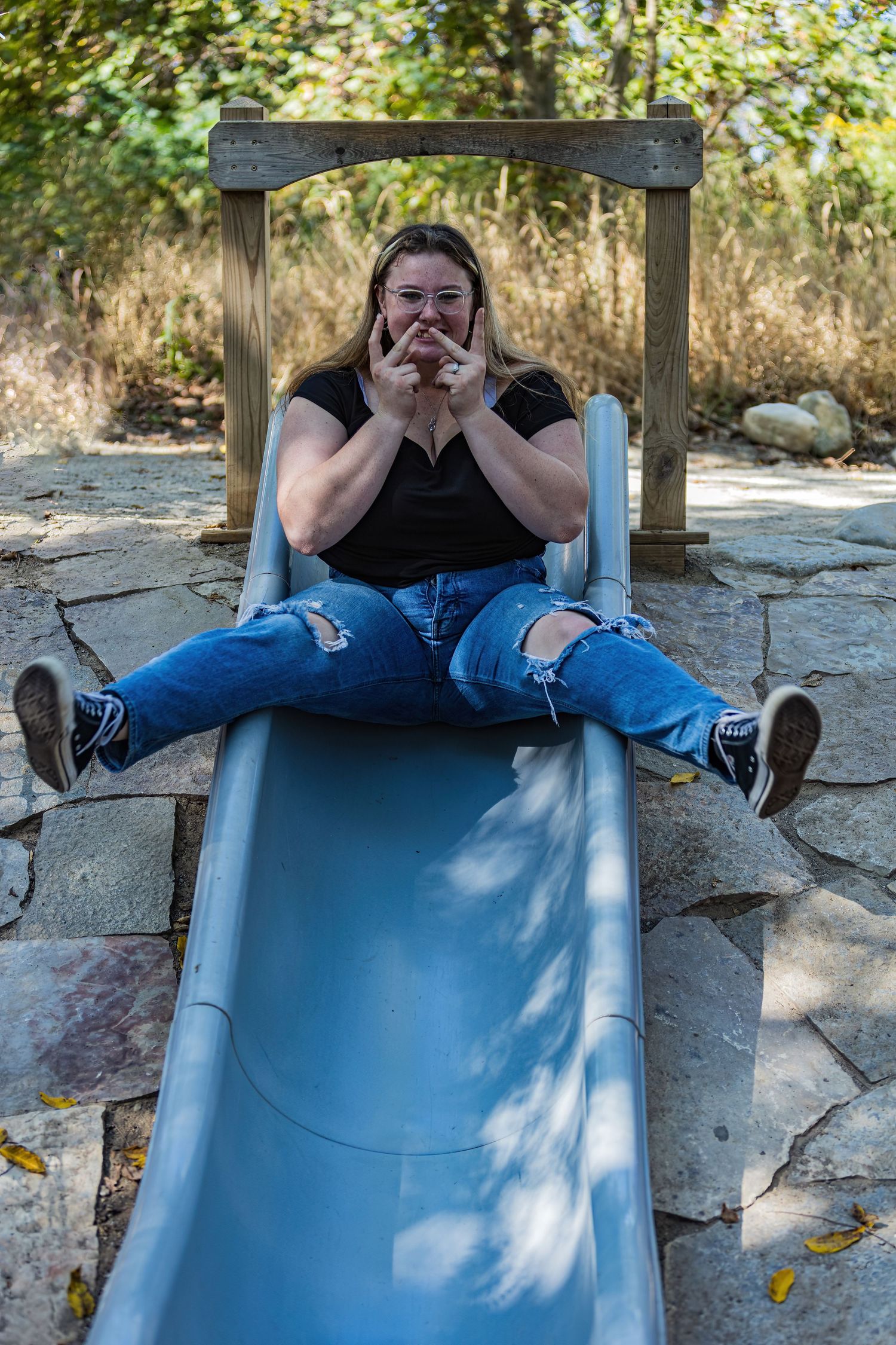 Person in black top and ripped jeans sits atop playground slide making animated expression.