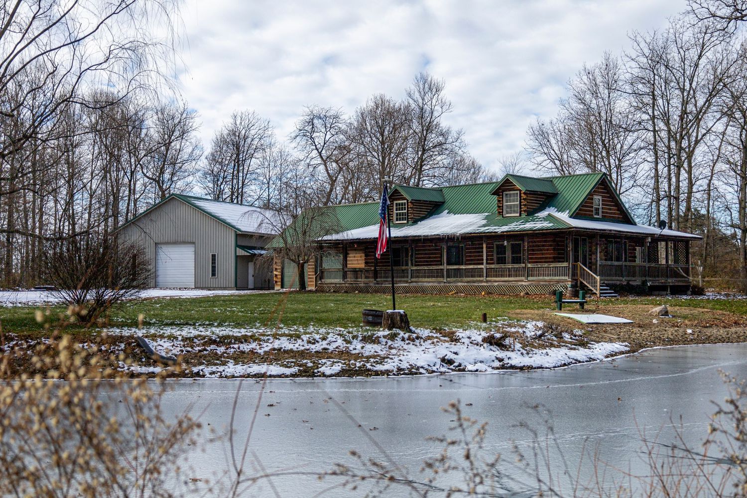 Log cabin home with green metal roof overlooking a frozen pond with detached garage.