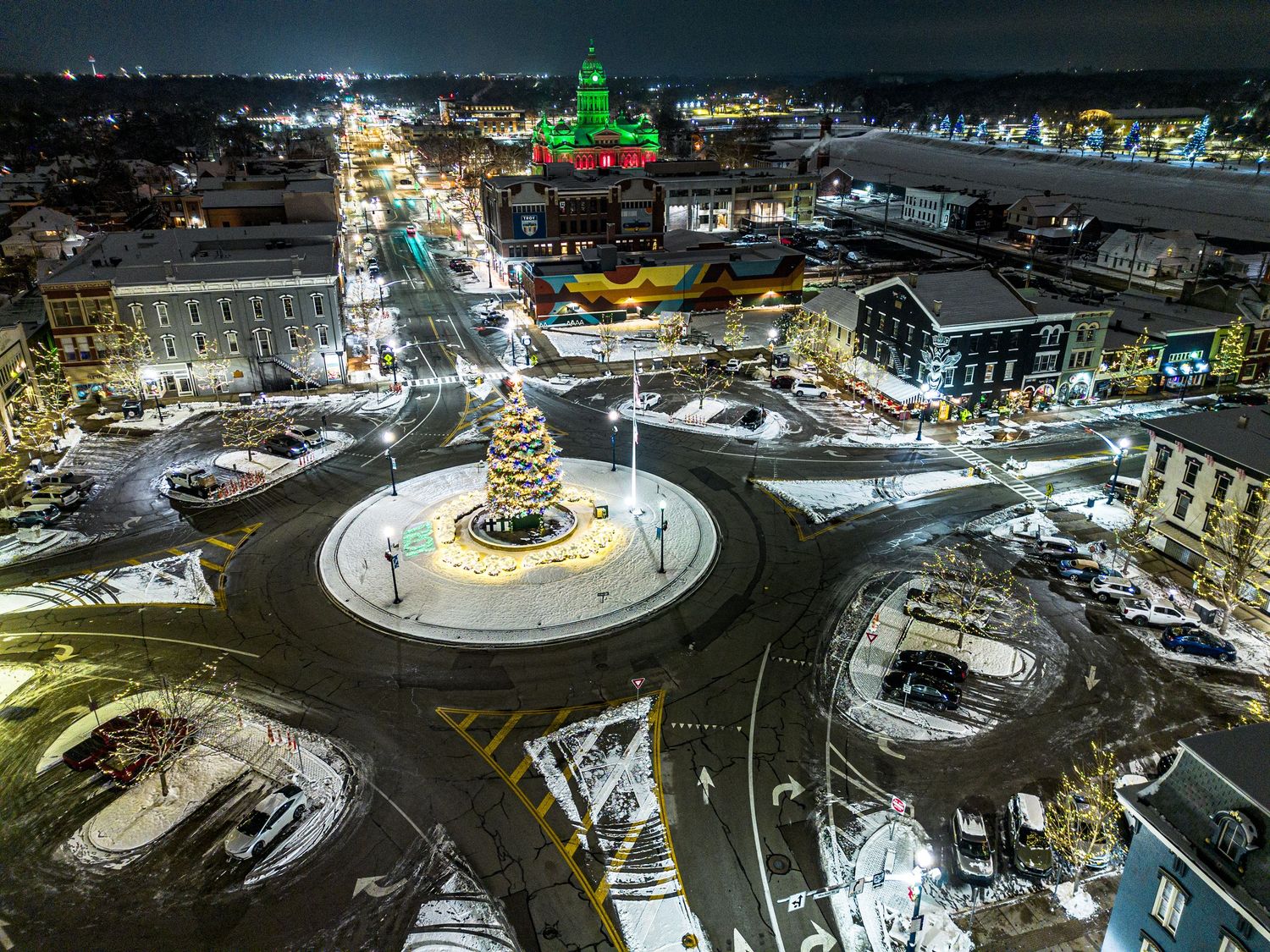 Nighttime aerial view of an illuminated roundabout with green holiday lighting and traffic.