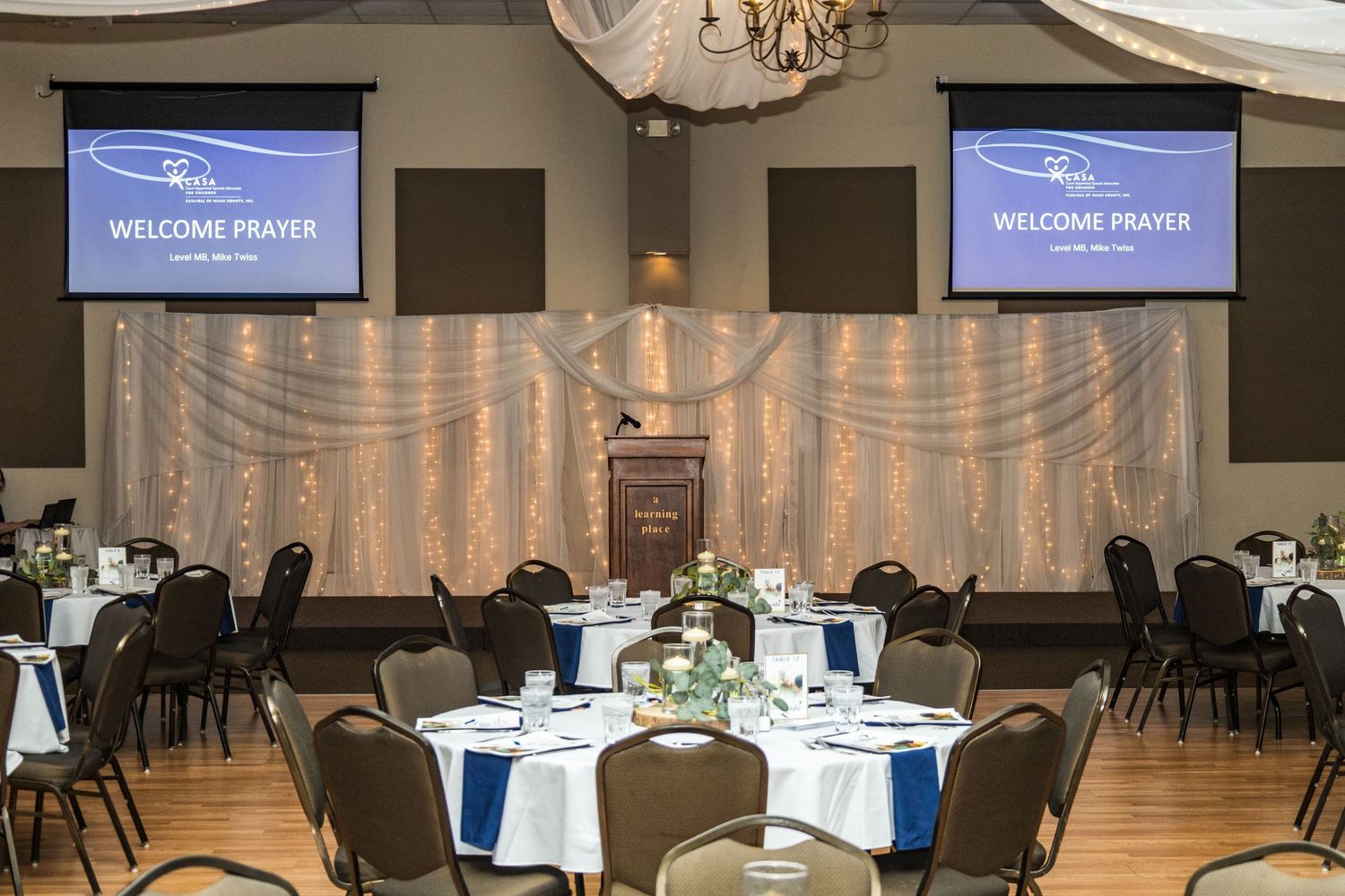 Modern event venue interior with multiple round tables set up for dining and wall-mounted displays.