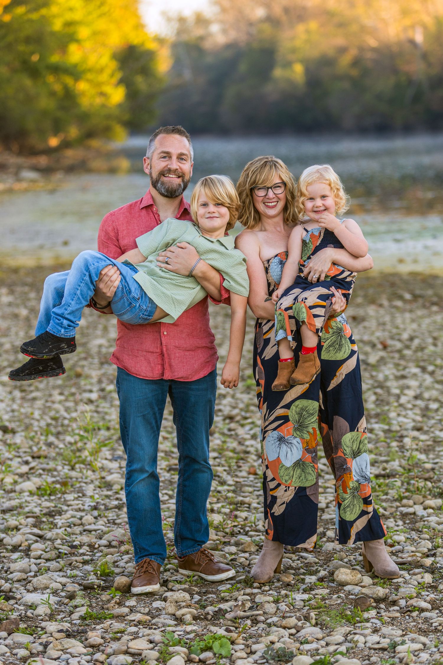 A family poses playfully together on a rocky riverbank during golden hour sunset.