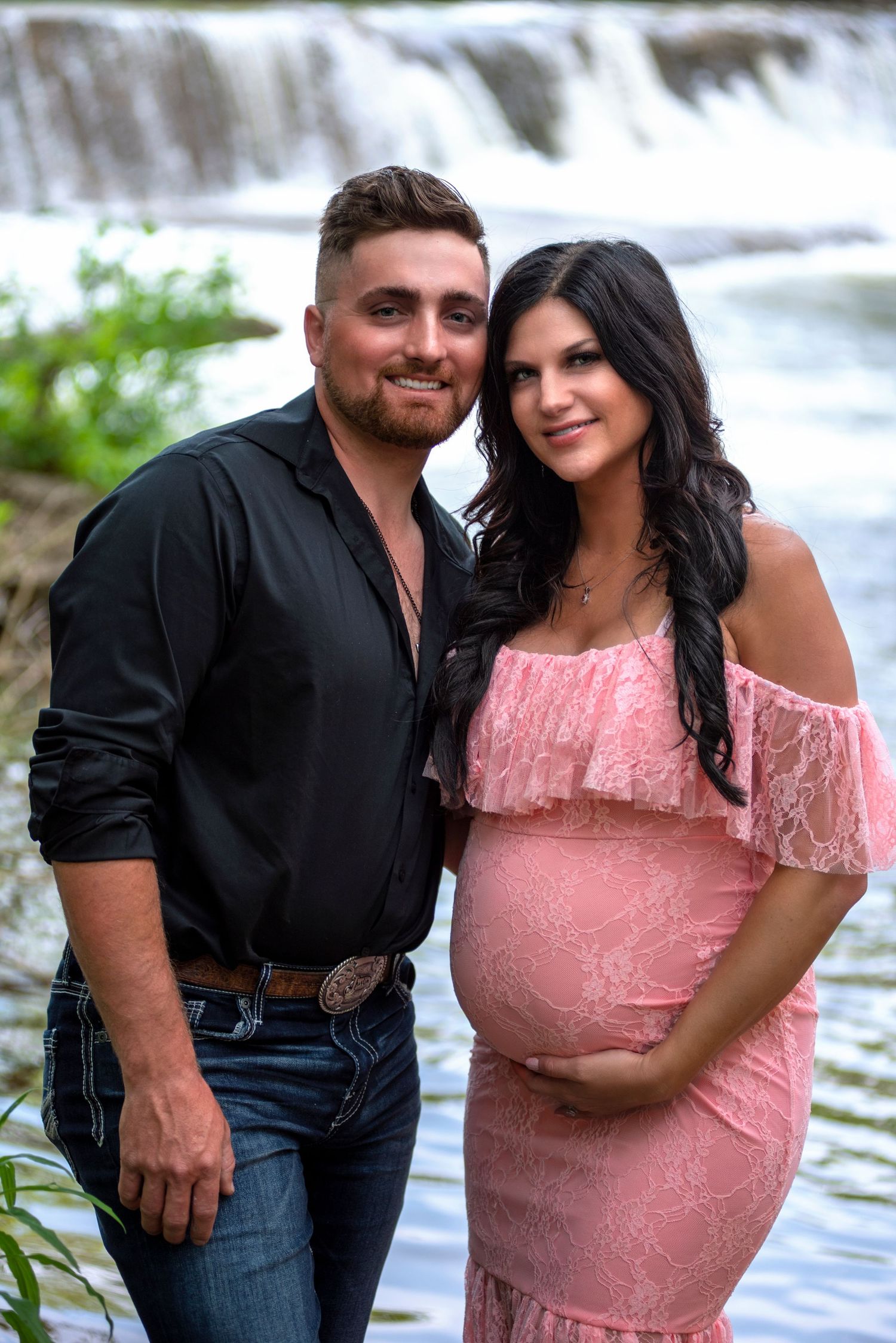 A couple poses together near a waterfall with her wearing a pink off-shoulder maternity dress.