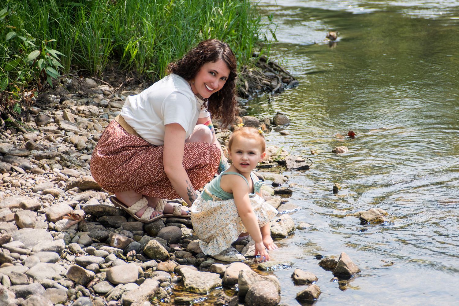 A parent and child feed ducks while sitting on rocks at the edge of a pond.