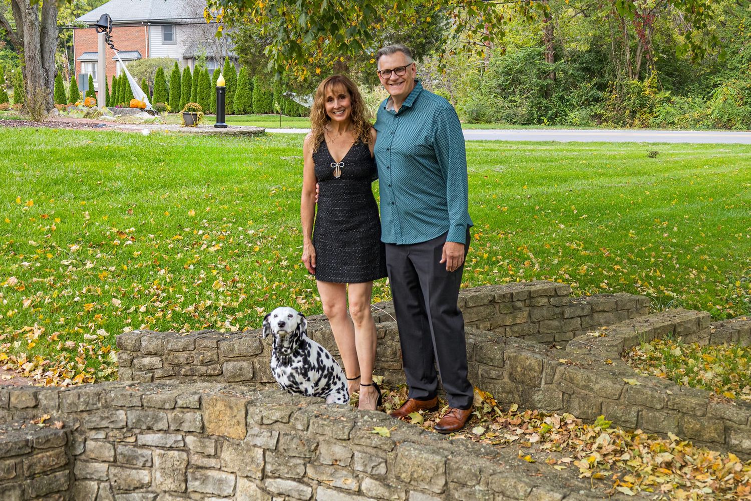 Two people stand together on a stone wall surrounded by autumn leaves and grass.