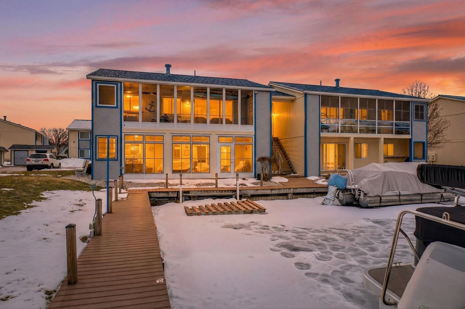 Modern waterfront home with warm interior lighting glowing at sunset against snowy landscape and wooden boardwalk.
