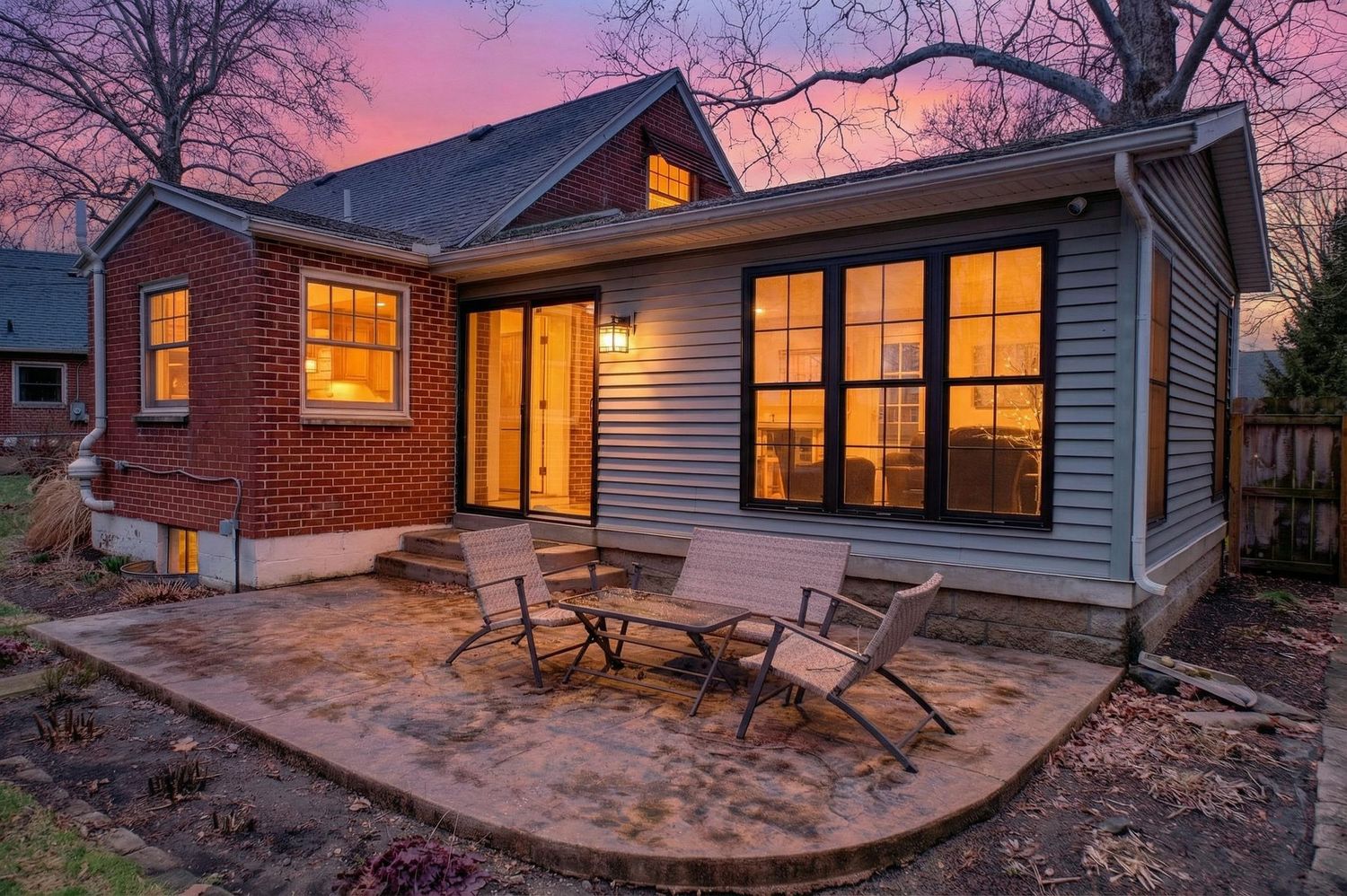 Modern ranch house exterior glows warmly at dusk with illuminated windows and curved patio under pink and purple sky.