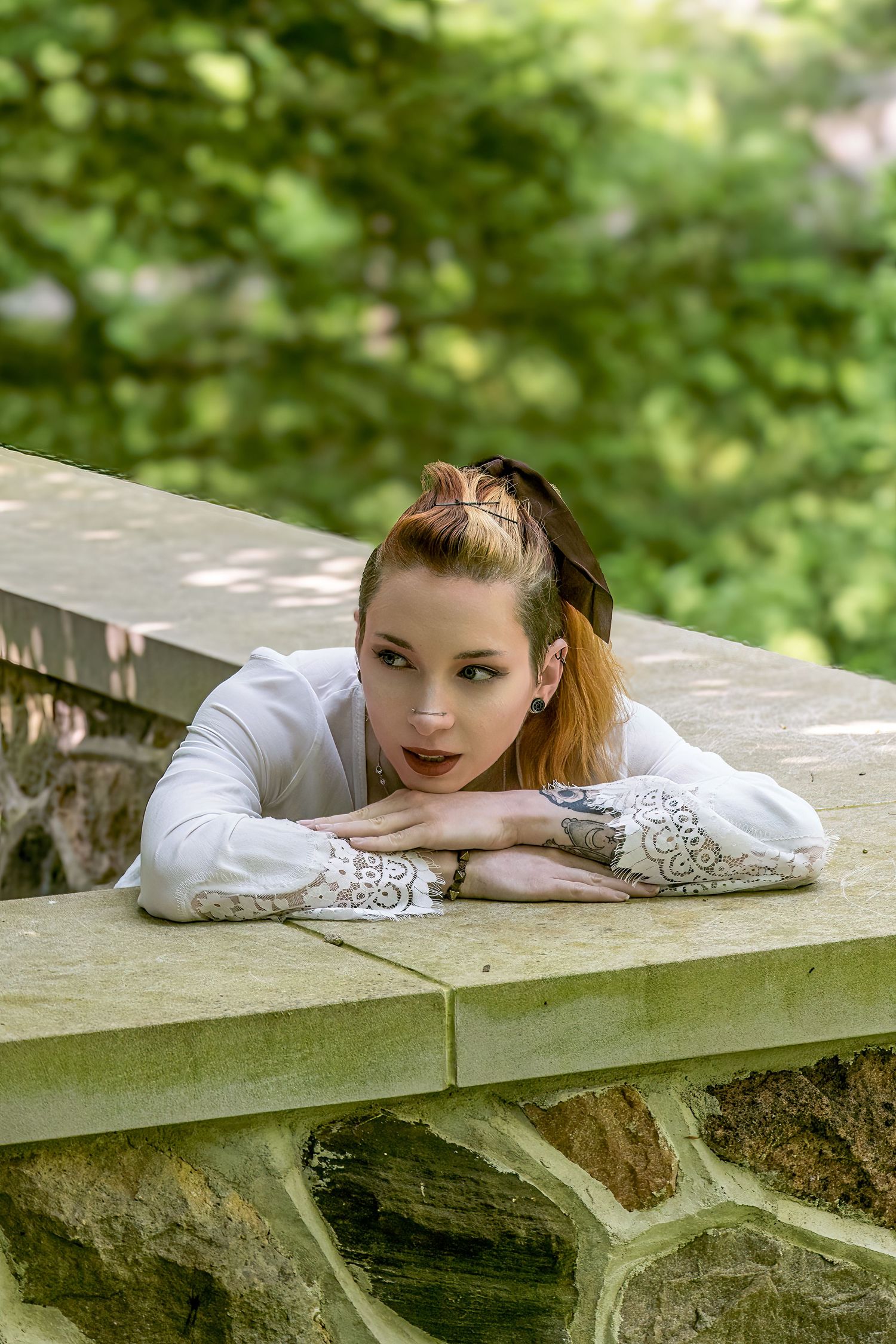 Portrait of a young woman in white lace dress resting on a stone wall outdoors.