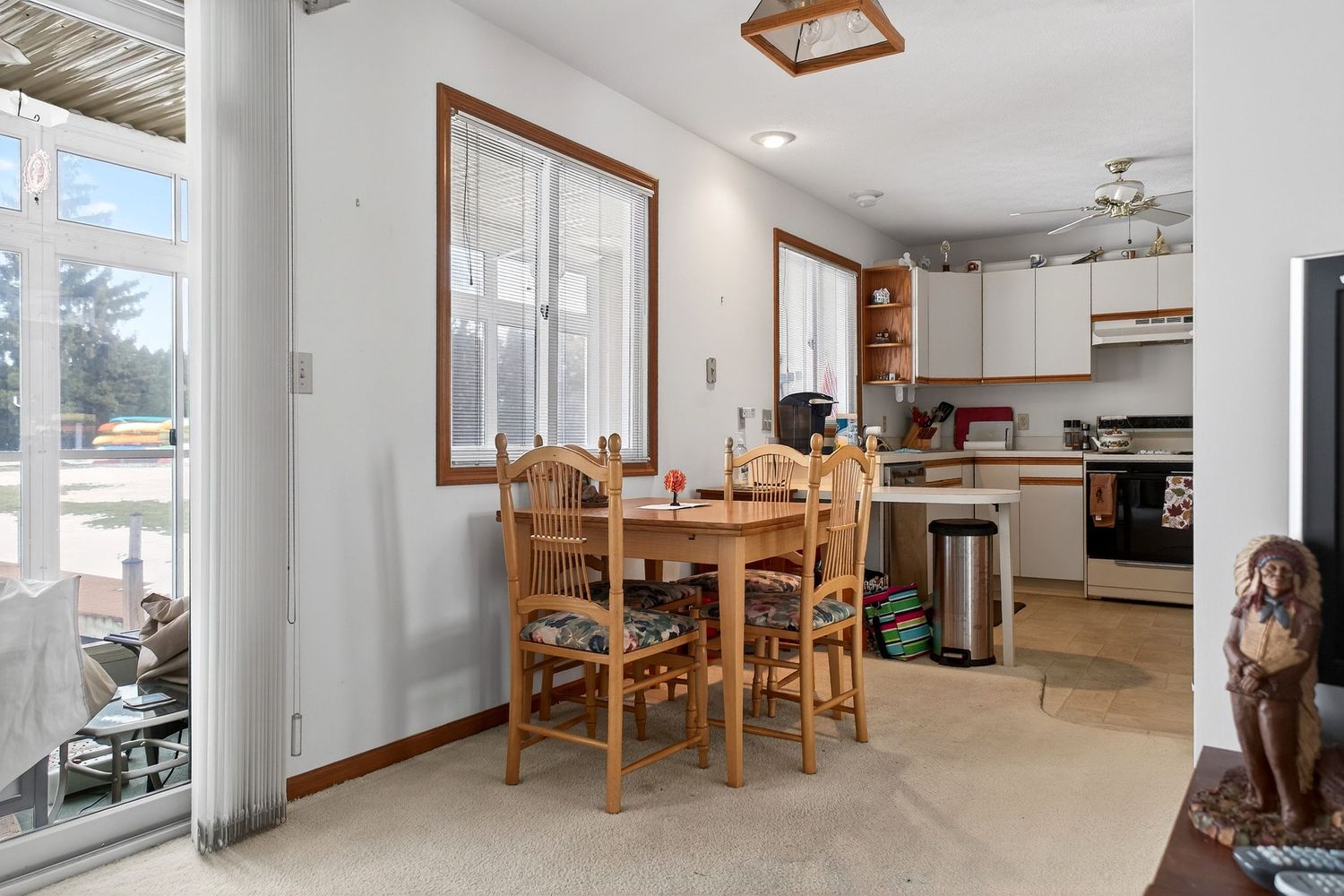Bright dining area with wooden table and chairs next to kitchen in mobile home with large windows and tile flooring.