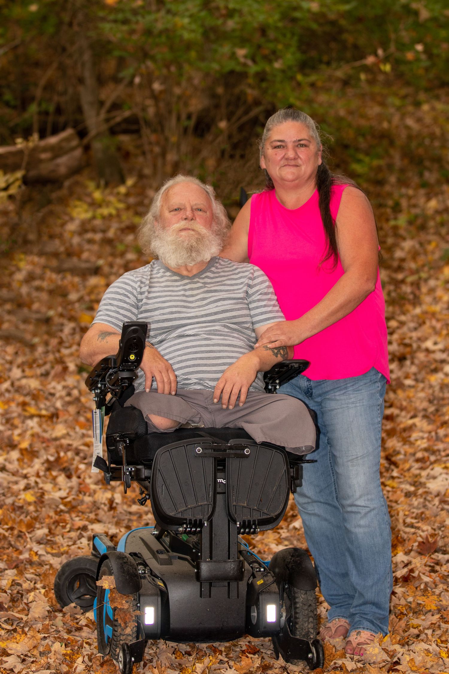 Person in motorized wheelchair and companion in pink shirt pose together among fallen autumn leaves.
