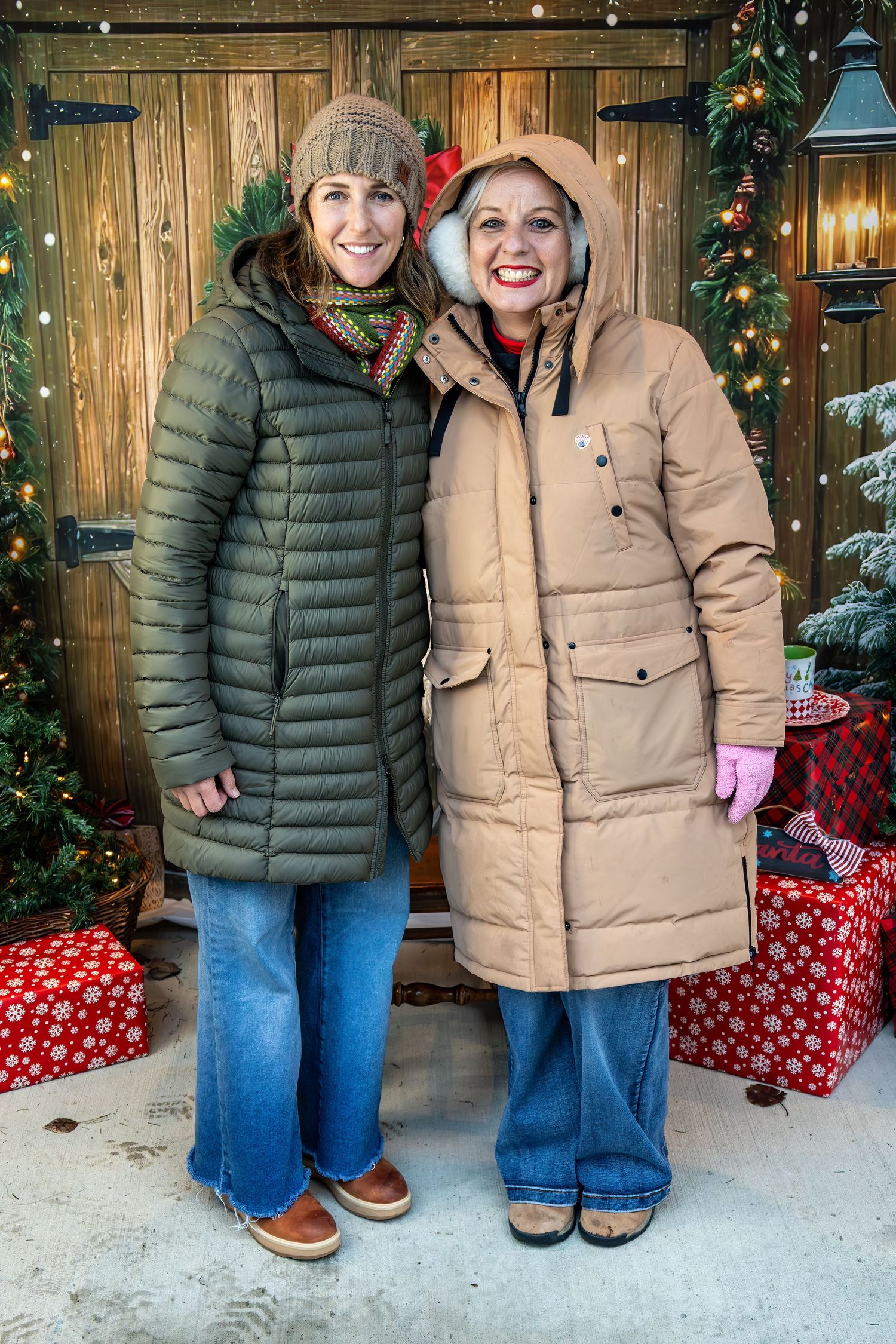 Two people in winter coats pose in front of a rustic wooden wall decorated with Christmas lights and wrapped presents.