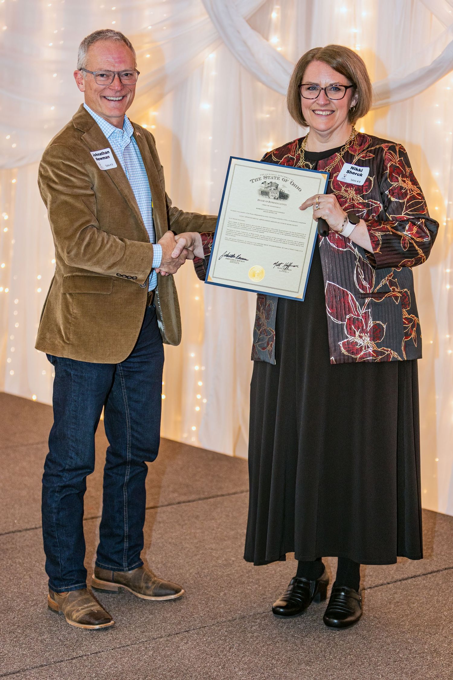 Two professionals pose with a certificate at an awards ceremony with twinkle lights in background.