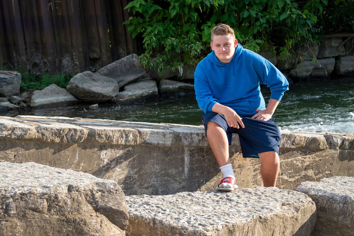 A person in a blue sweater poses on rocky formations near water.