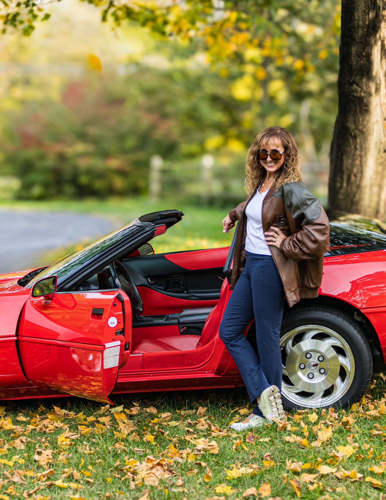 A bright red sports car sits parked on fallen autumn leaves in a scenic outdoor setting.