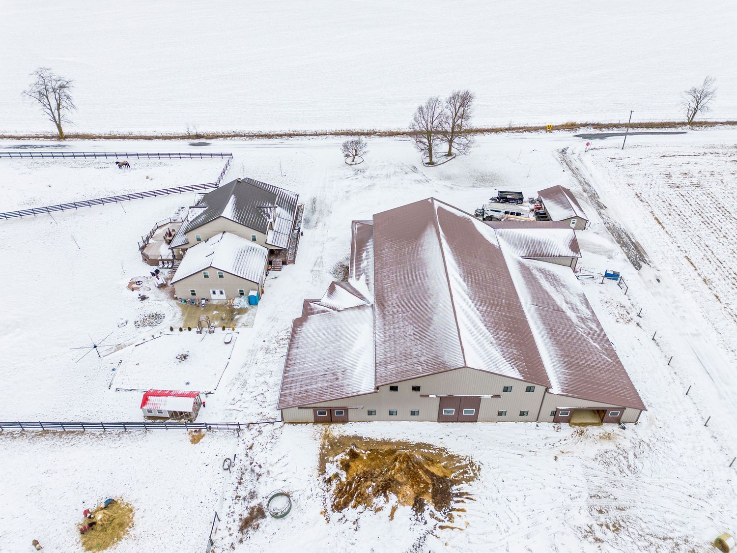 Aerial winter view of snow covered farm buildings and barns surrounded by white fields and scattered trees.