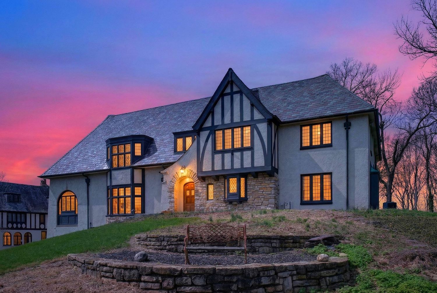 Tudor-style luxury home with illuminated windows against a dramatic purple and pink sunset sky.