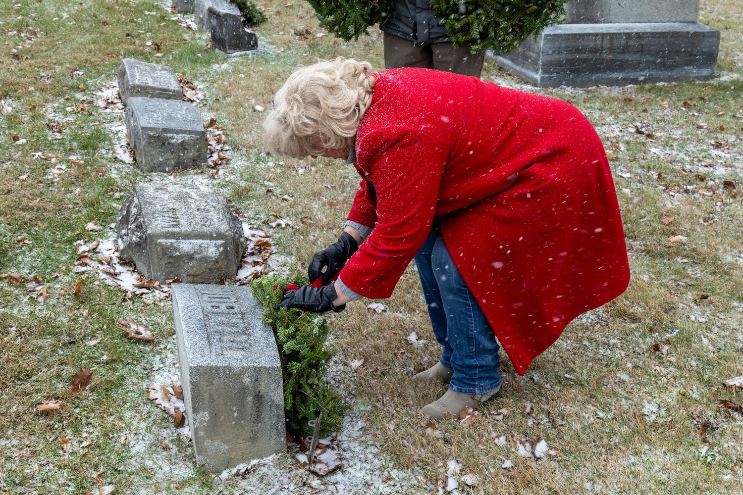 Person in red coat placing a wreath on a gravestone during winter memorial service.