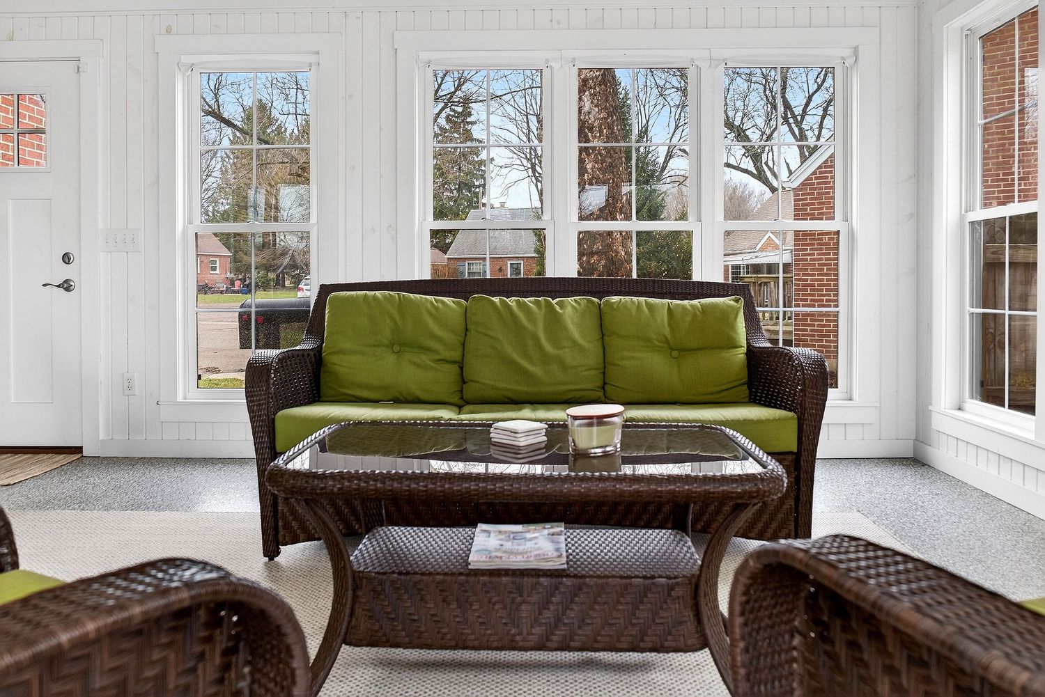 Bright sunroom with olive green cushioned wicker furniture and large windows overlooking winter trees.