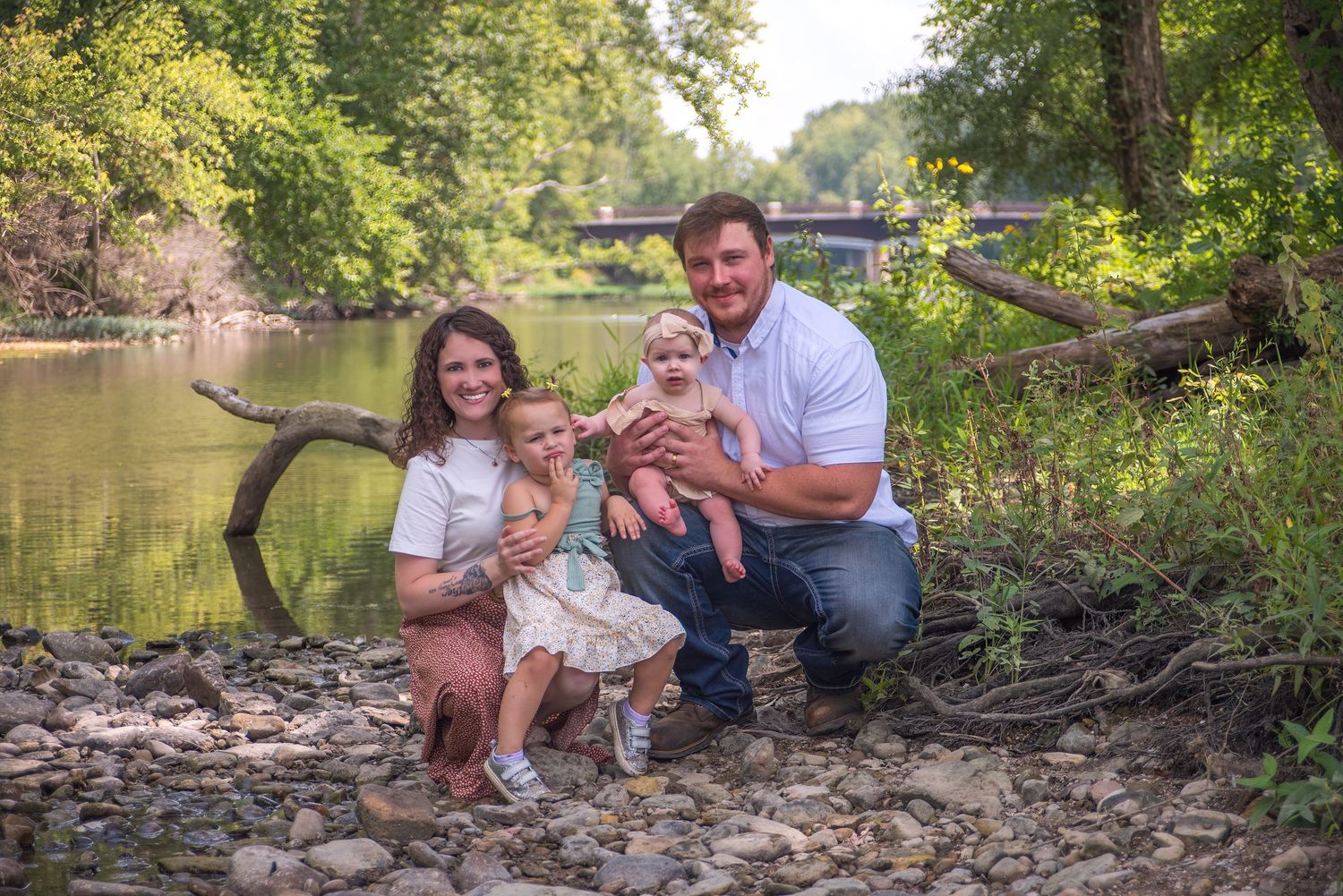 A family sits together on a fallen tree branch by a peaceful river on a sunny day.