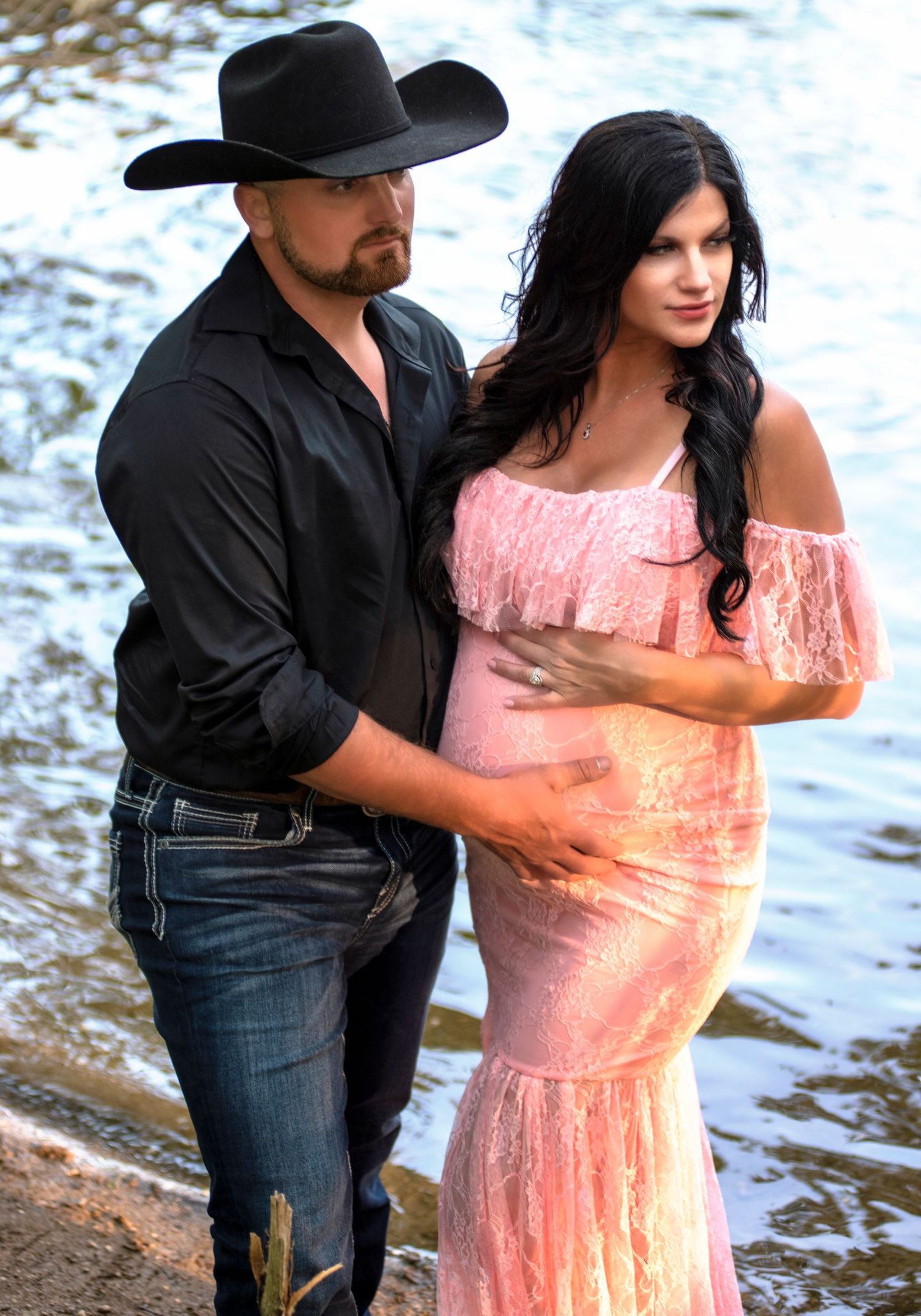 A couple poses by water, with him in a black cowboy hat and her in a pink flowing dress.