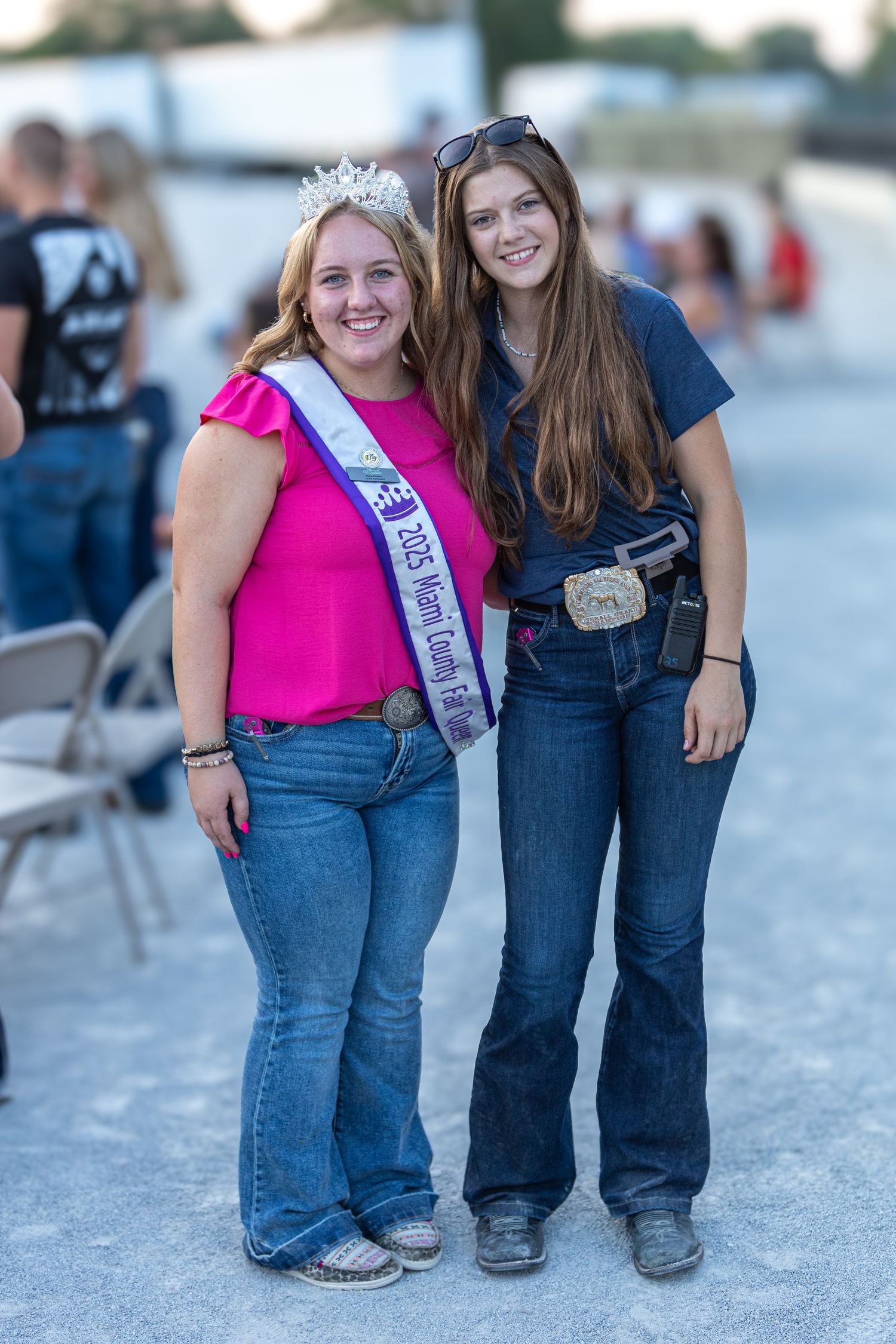 Two people wearing pageant sashes pose together on concrete ground.