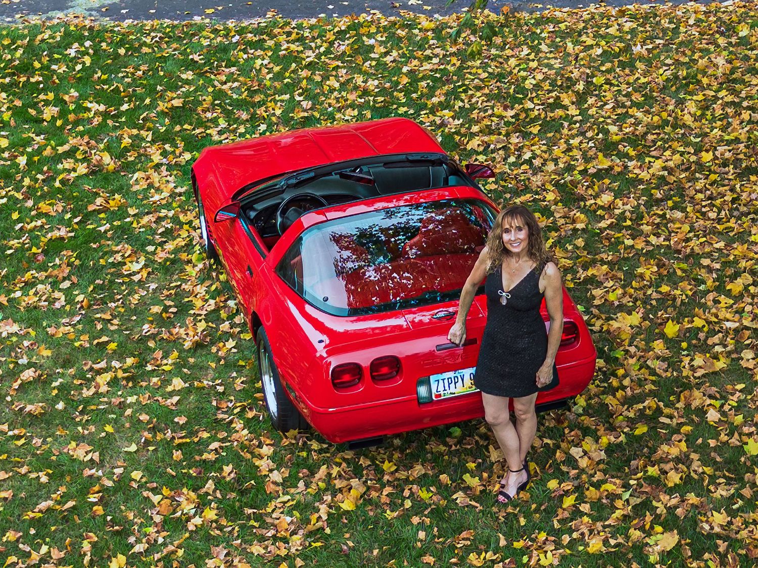 A red sports car parked on fallen autumn leaves with scenic fall foliage in the background.
