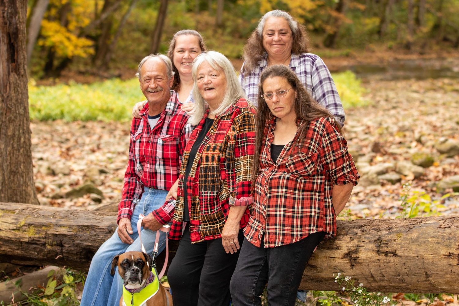 A group in matching plaid shirts poses together on a fallen log in an autumn forest setting.