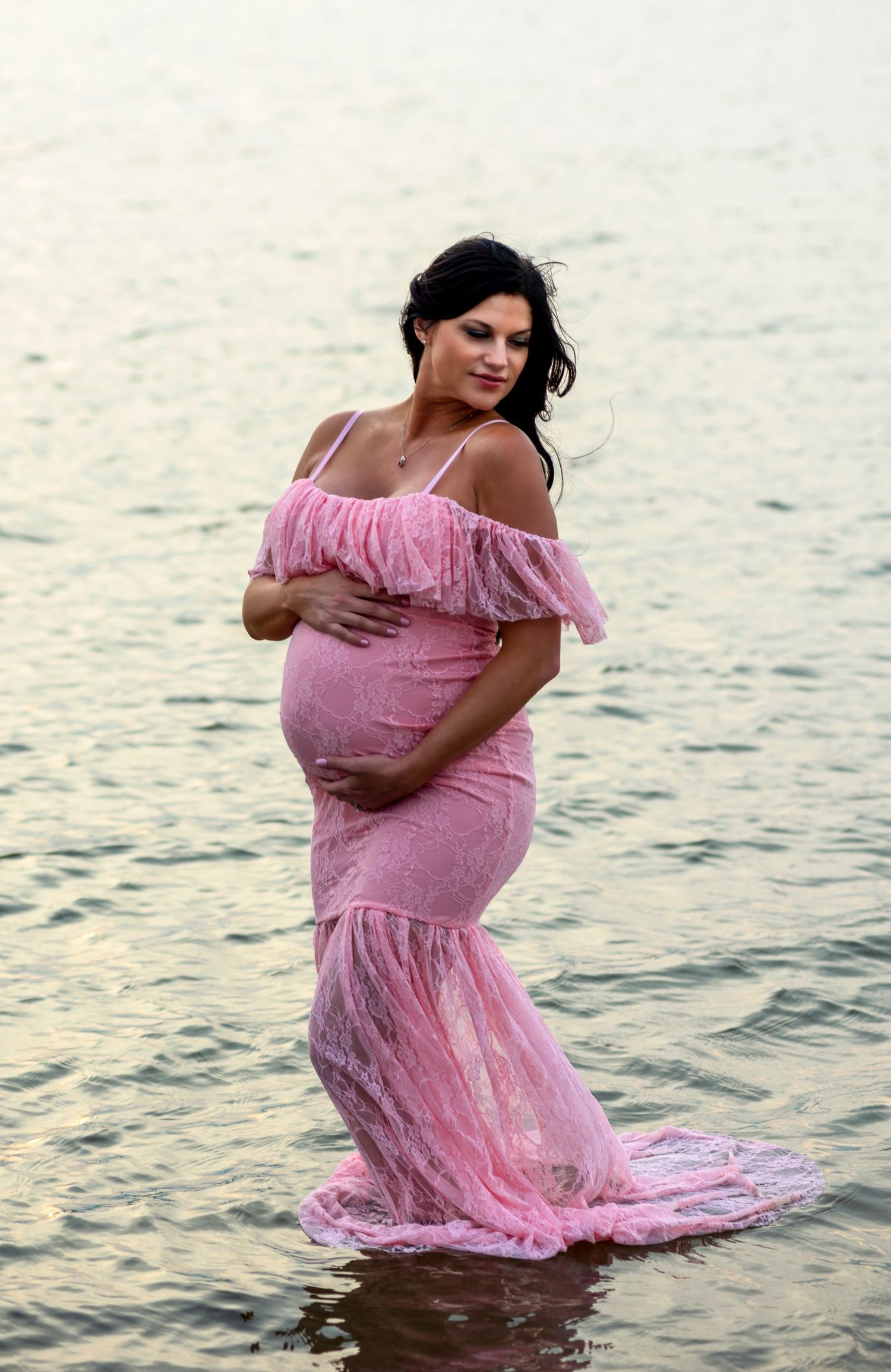 A maternity photo of someone in a pink ruffled dress standing in shallow water at sunset.