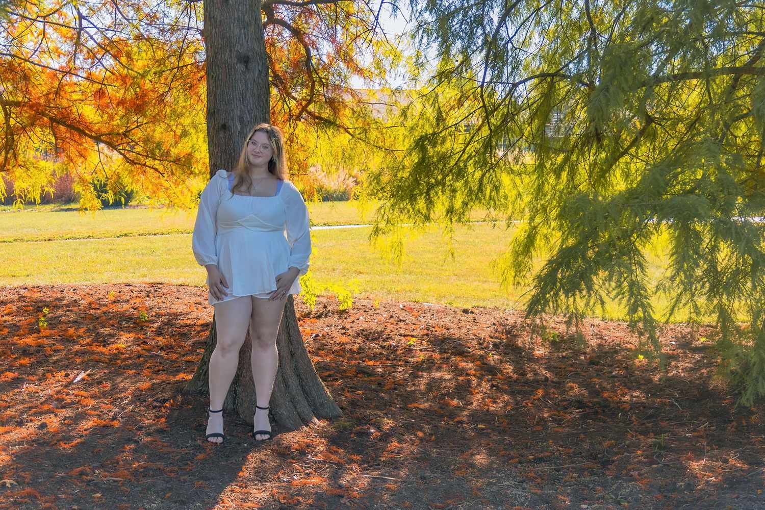 Someone in a white sweater poses against a tree with autumn foliage in the background.