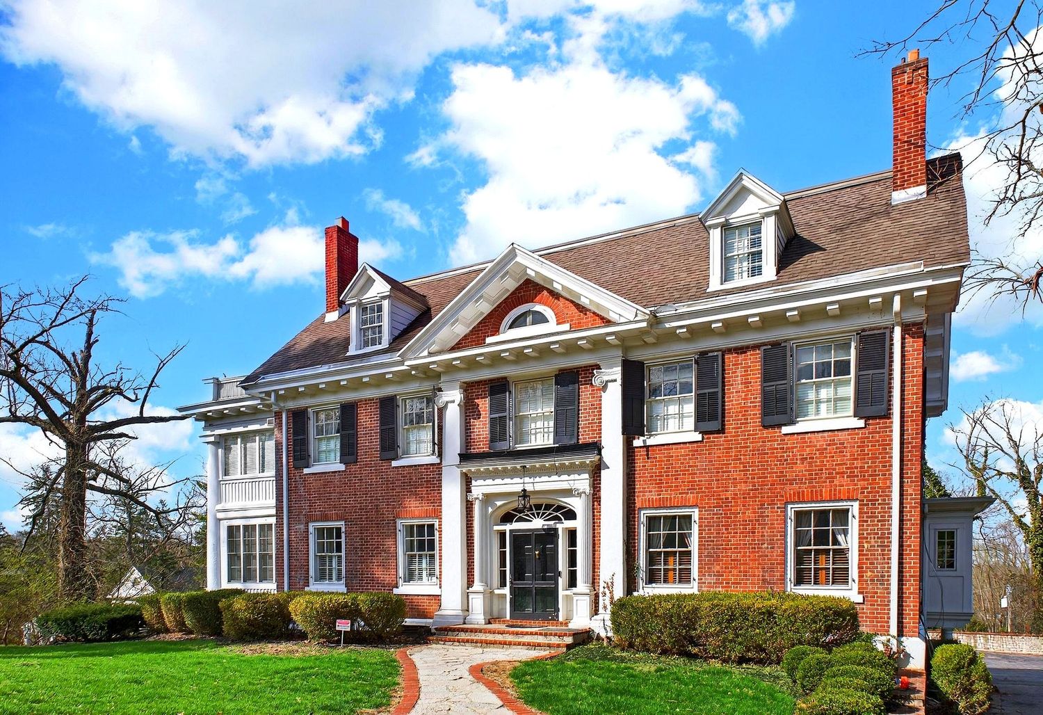 Stately red brick Georgian colonial mansion with white columns and black shutters under bright blue sky.