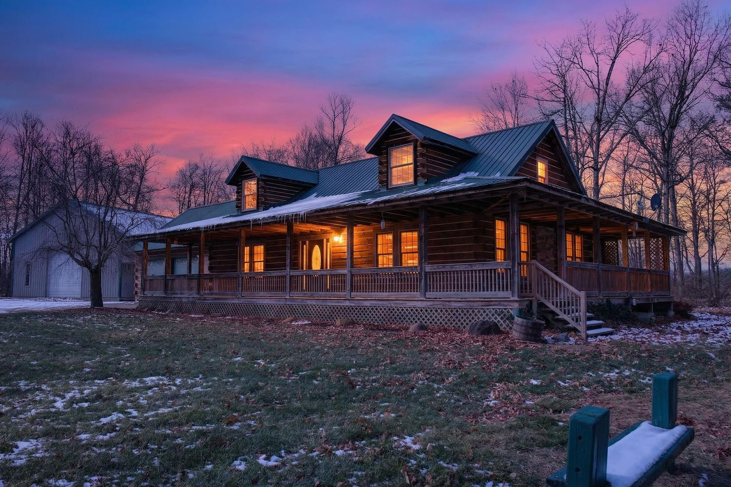 A rustic log cabin glows warmly against a dramatic purple and orange sunset sky with bare winter trees in the background.