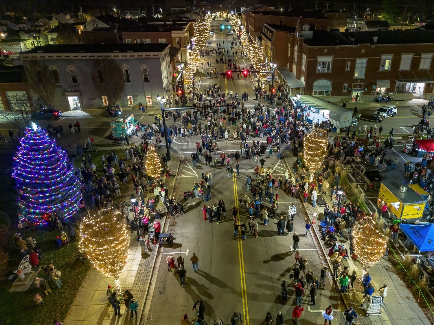 Night view of a holiday celebration with blue and gold lit trees surrounded by festival crowds.