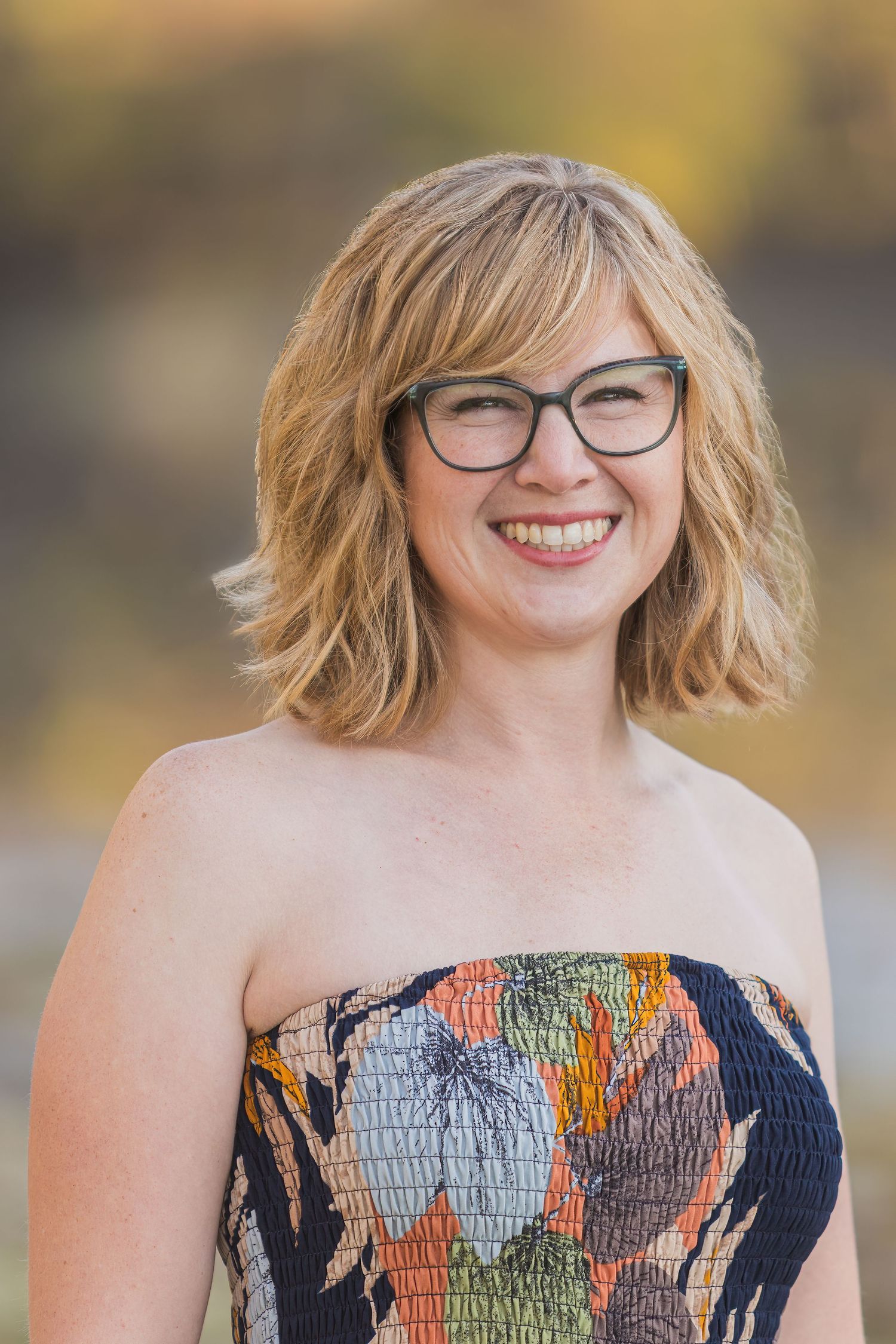 Portrait of someone wearing glasses and a patterned strapless top against a warm autumn background.