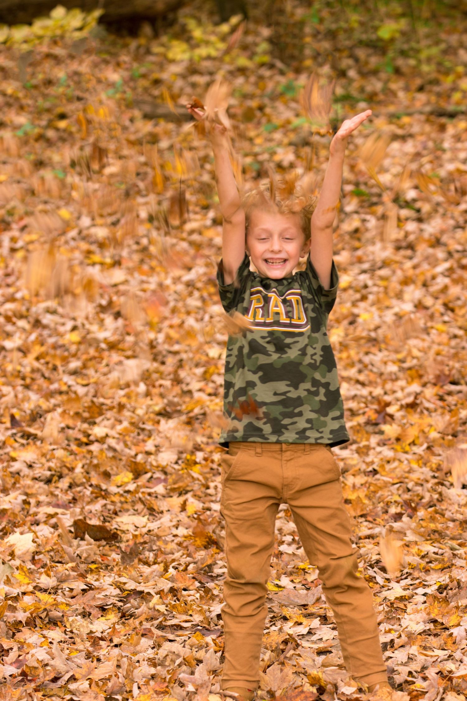Child in camouflage shirt throws autumn leaves in the air with excitement.