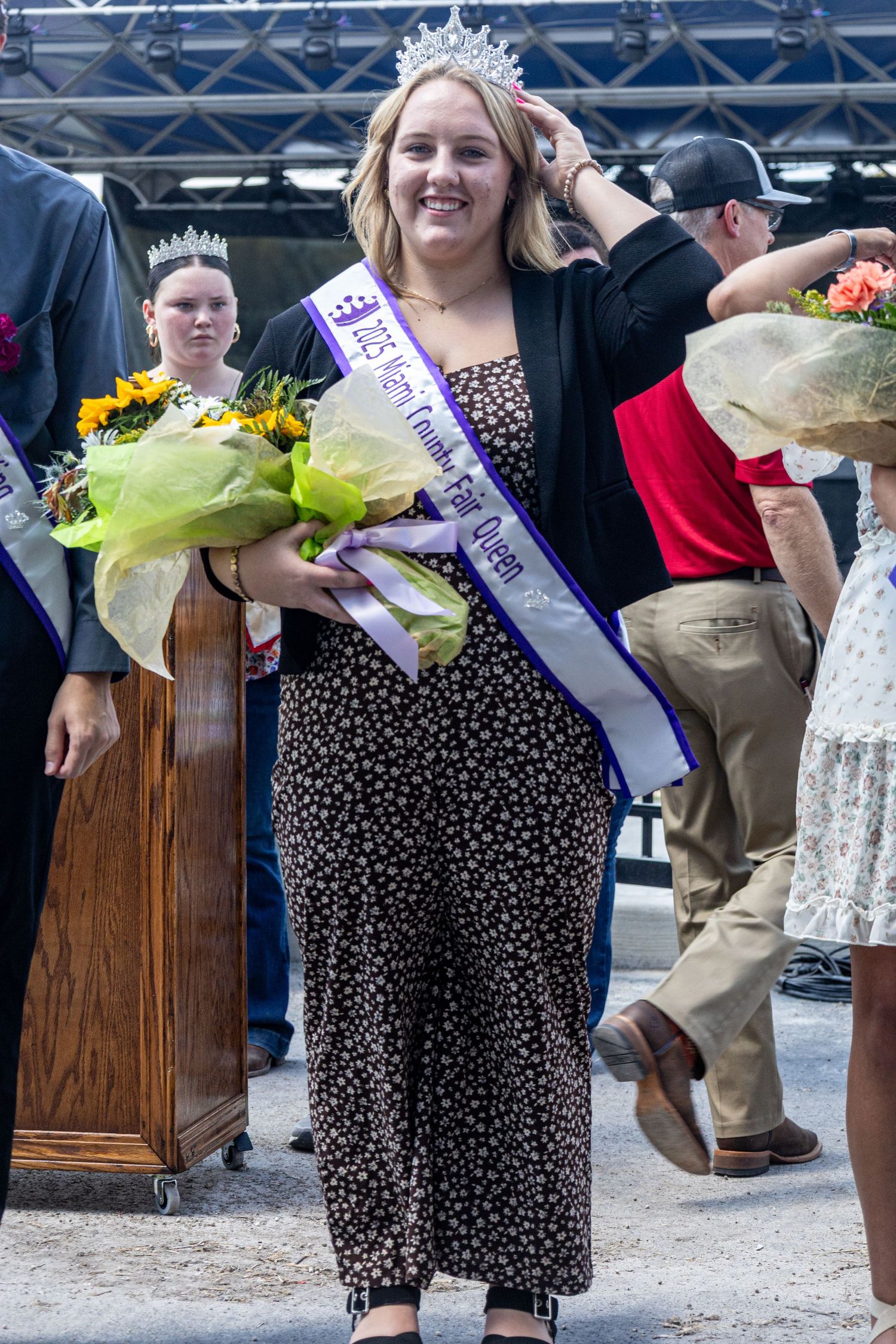 Group wearing sashes and holding flower bouquets stand in line at awards ceremony.