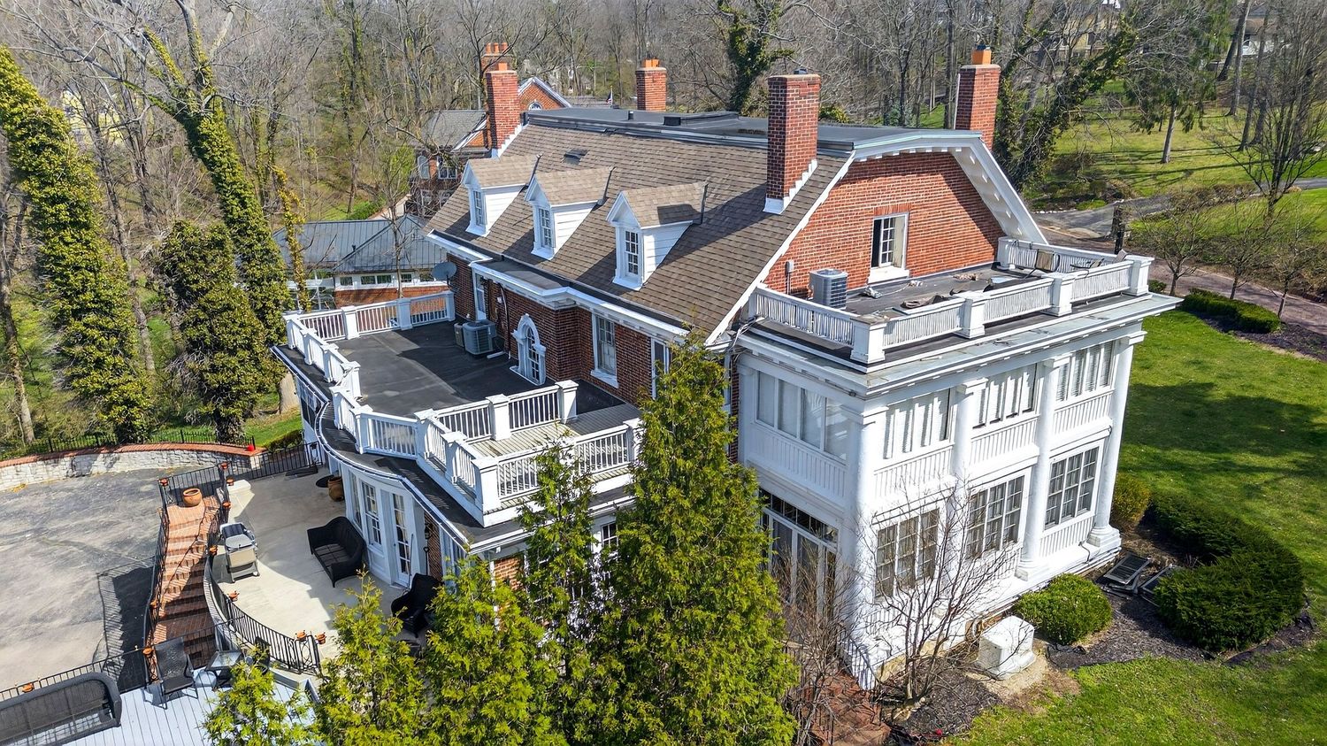 A large white colonial-style mansion with multiple chimneys and dormers is surrounded by trees in early spring.
