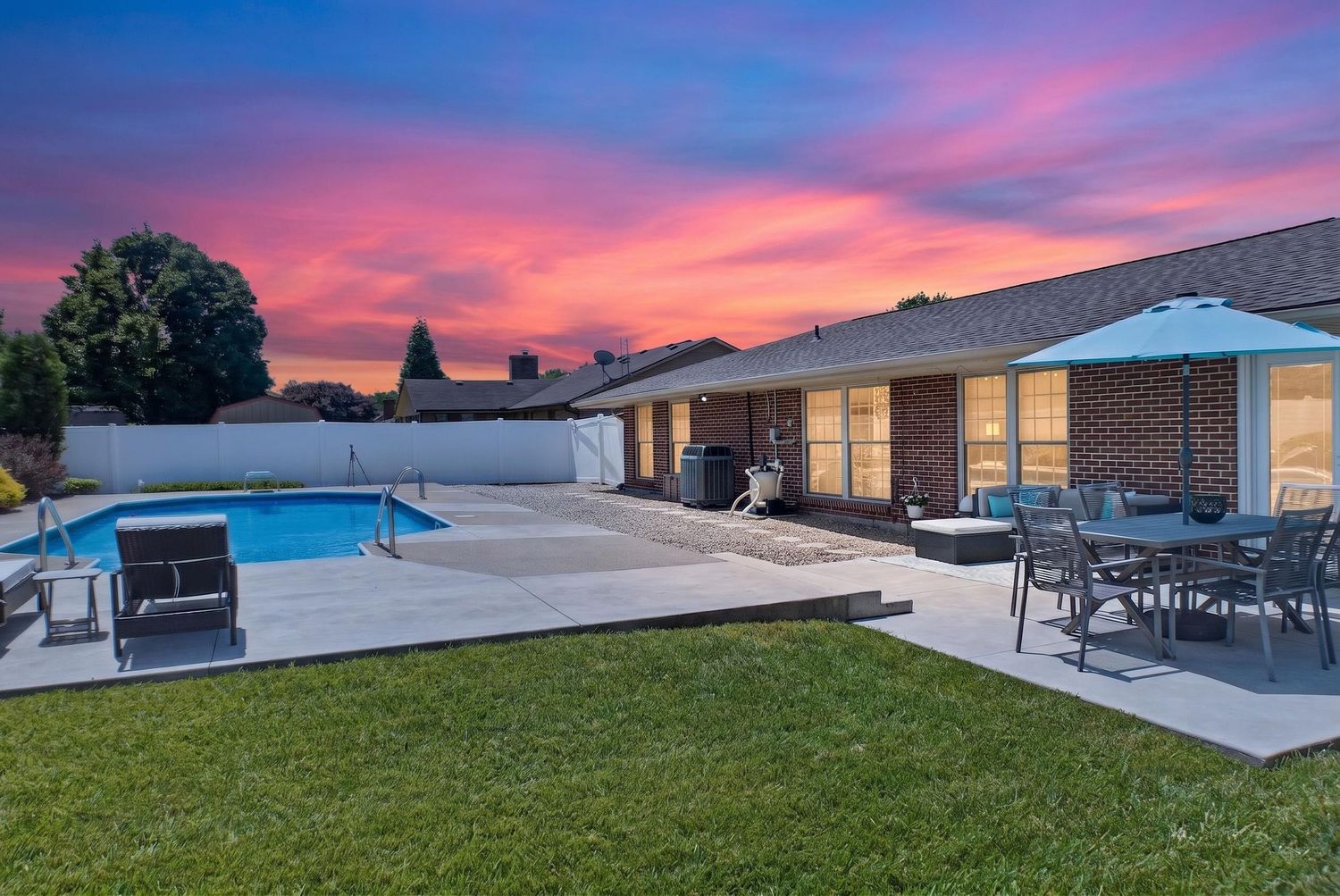 Backyard pool and patio with brick home, outdoor dining set, and vibrant pink and purple sunset sky.