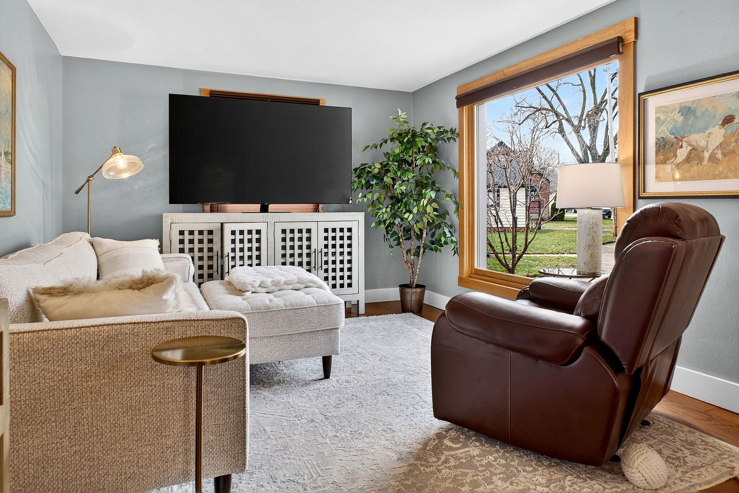 Modern living room interior with brown leather sofa, gray walls, hardwood floors and large windows letting in natural light.