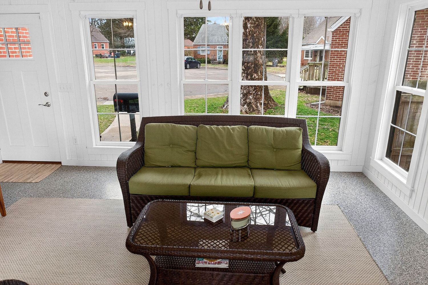 A bright sunroom with olive green couches, large windows, and a dark wood coffee table creates a cozy seating area.
